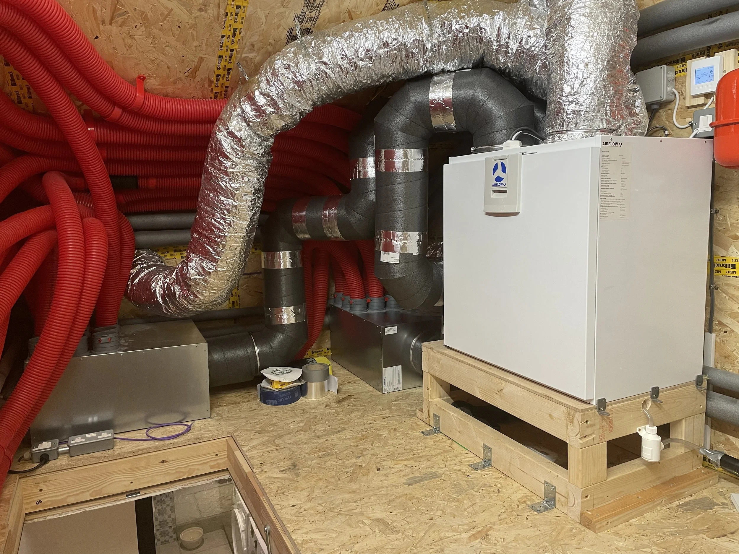 A room with insulation and mechanical equipment including red flexible tubing, black ductwork, and a white HVAC unit on a wooden platform.