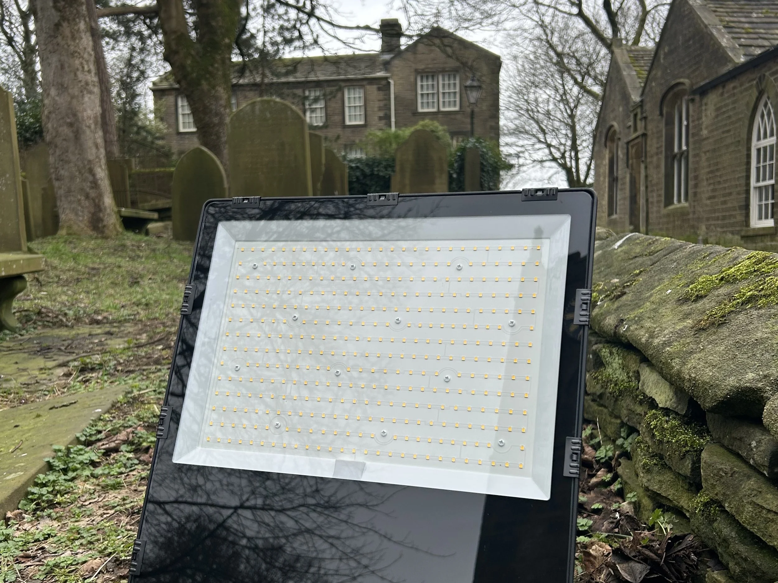 LED floodlight on a grassy ground in front of a stone wall, trees, and old brick houses with arched windows, during a cloudy day.