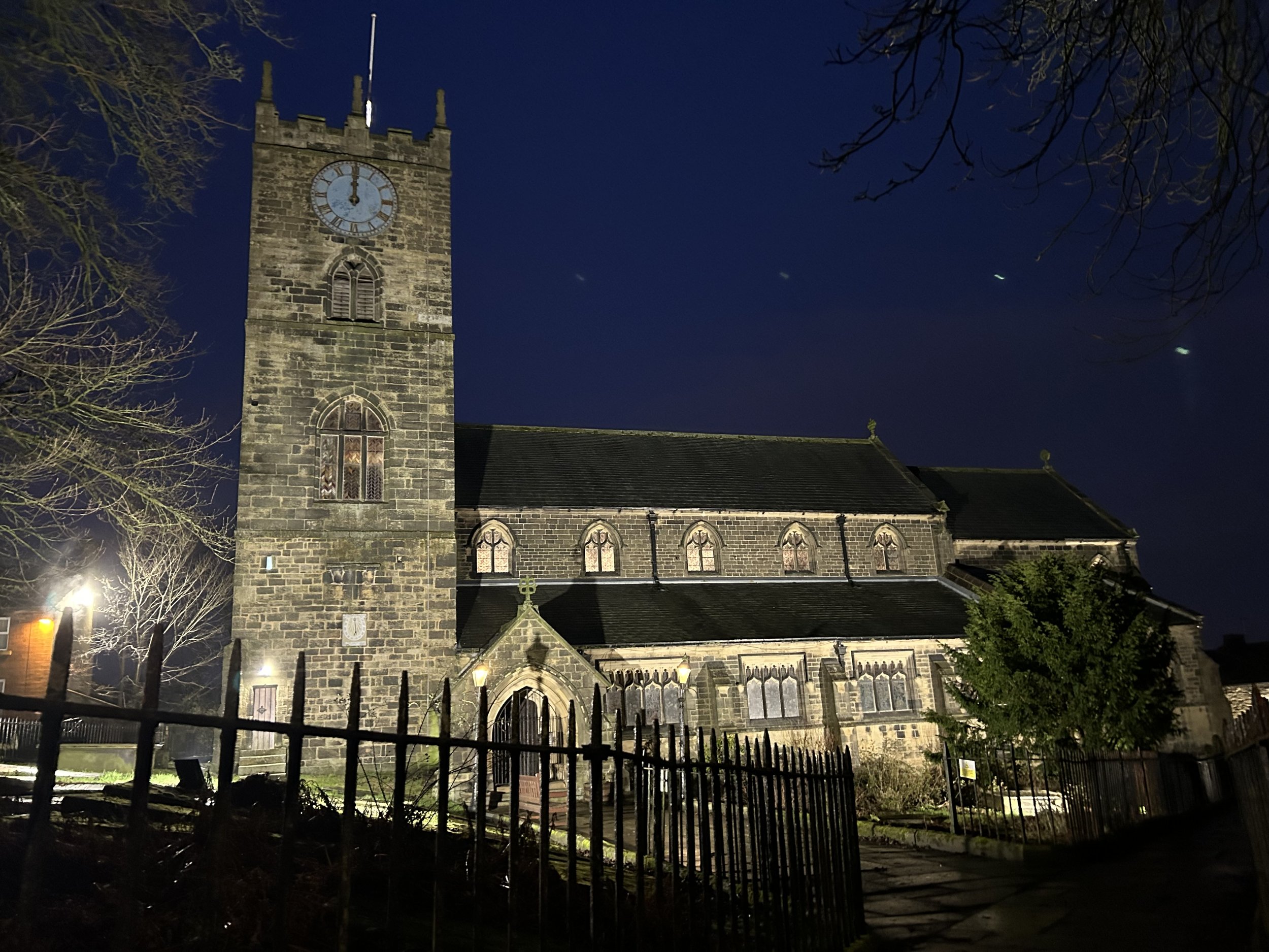 Nighttime view of St Michael & All Angel's Church in Haworth located next to the Brontë Parsonage Museum with a stone tower and clock, surrounded by bare trees and a black metal fence, illuminated by exterior lighting.