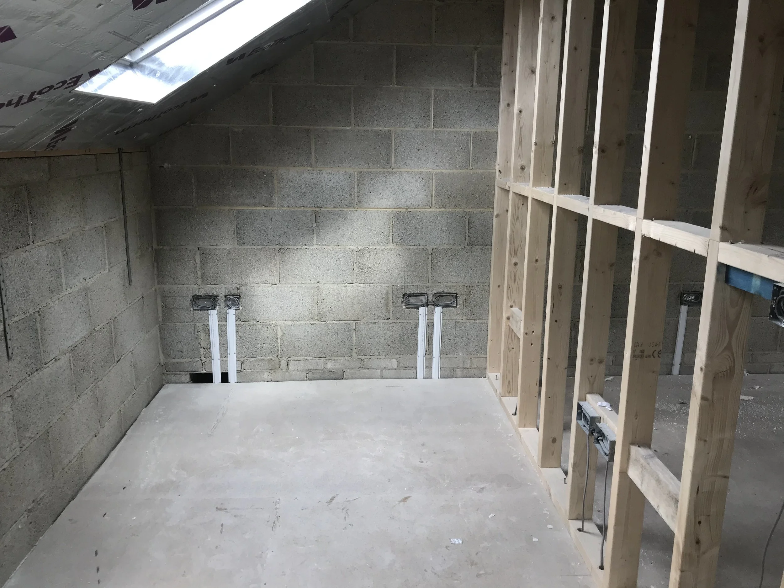 An unfinished attic space with concrete-block walls, exposed ceiling insulation, and a wooden frame wall with electrical wiring and outlets.