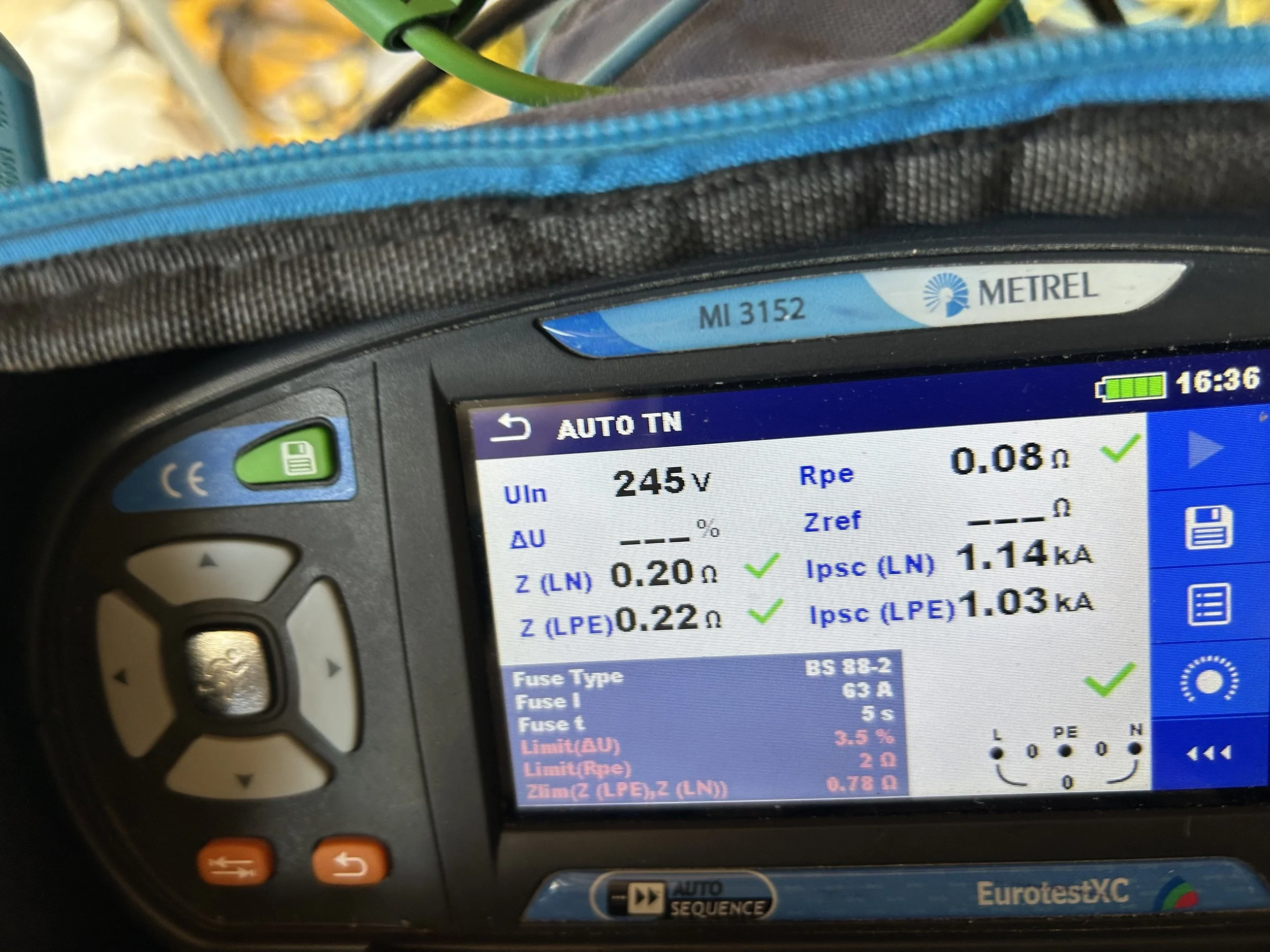 Close-up of a portable electronic device display showing electrical testing measurements, with a black and gray textured case, a green button, and an orange button nearby.