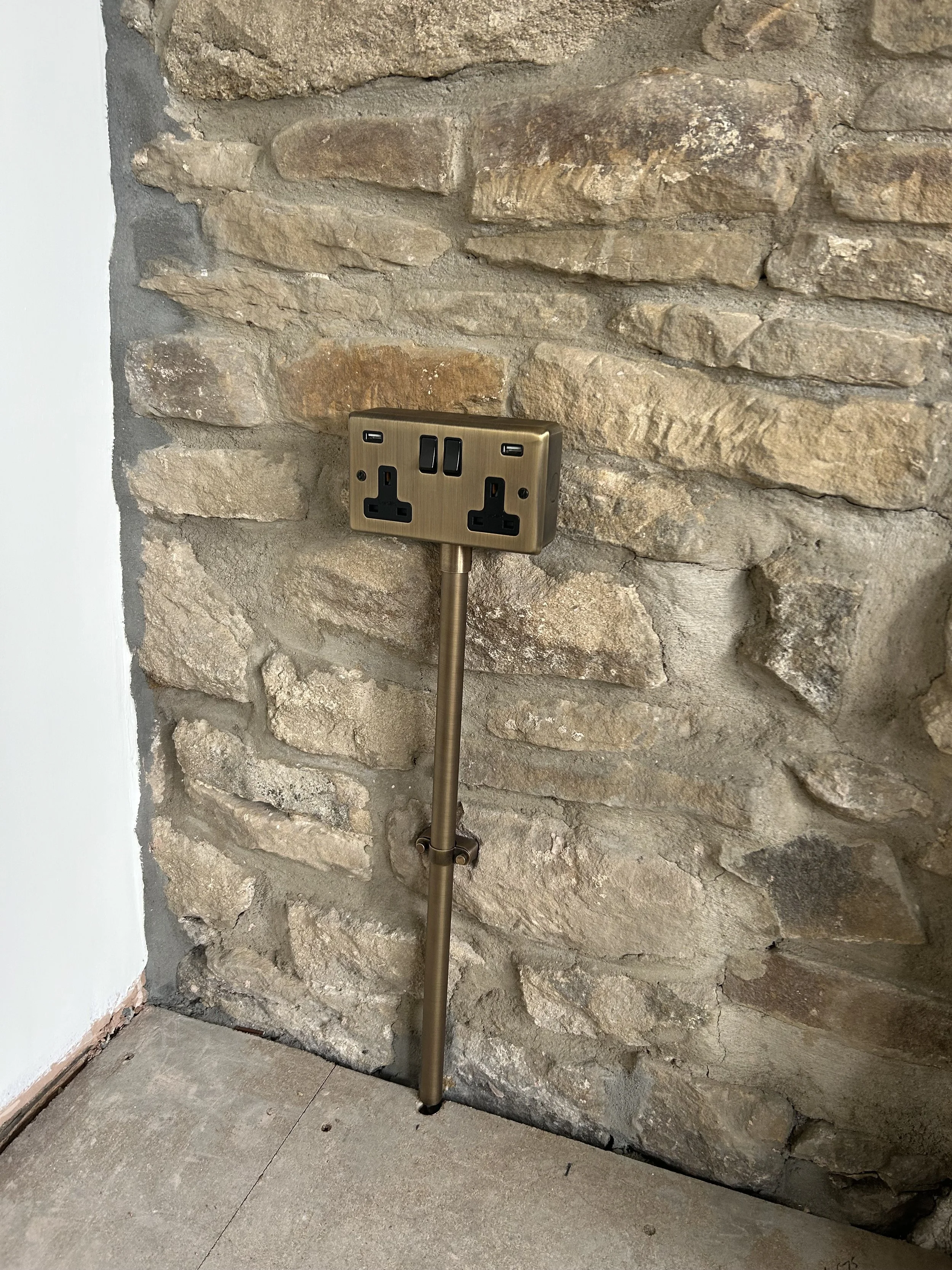 A power outlet with two UK-style sockets and two USB ports mounted on a bespoke antique brass-colored conduit & switch plate, against a rustic stone wall. The lower part of the wall is unfinished, with gray and white painted sections.