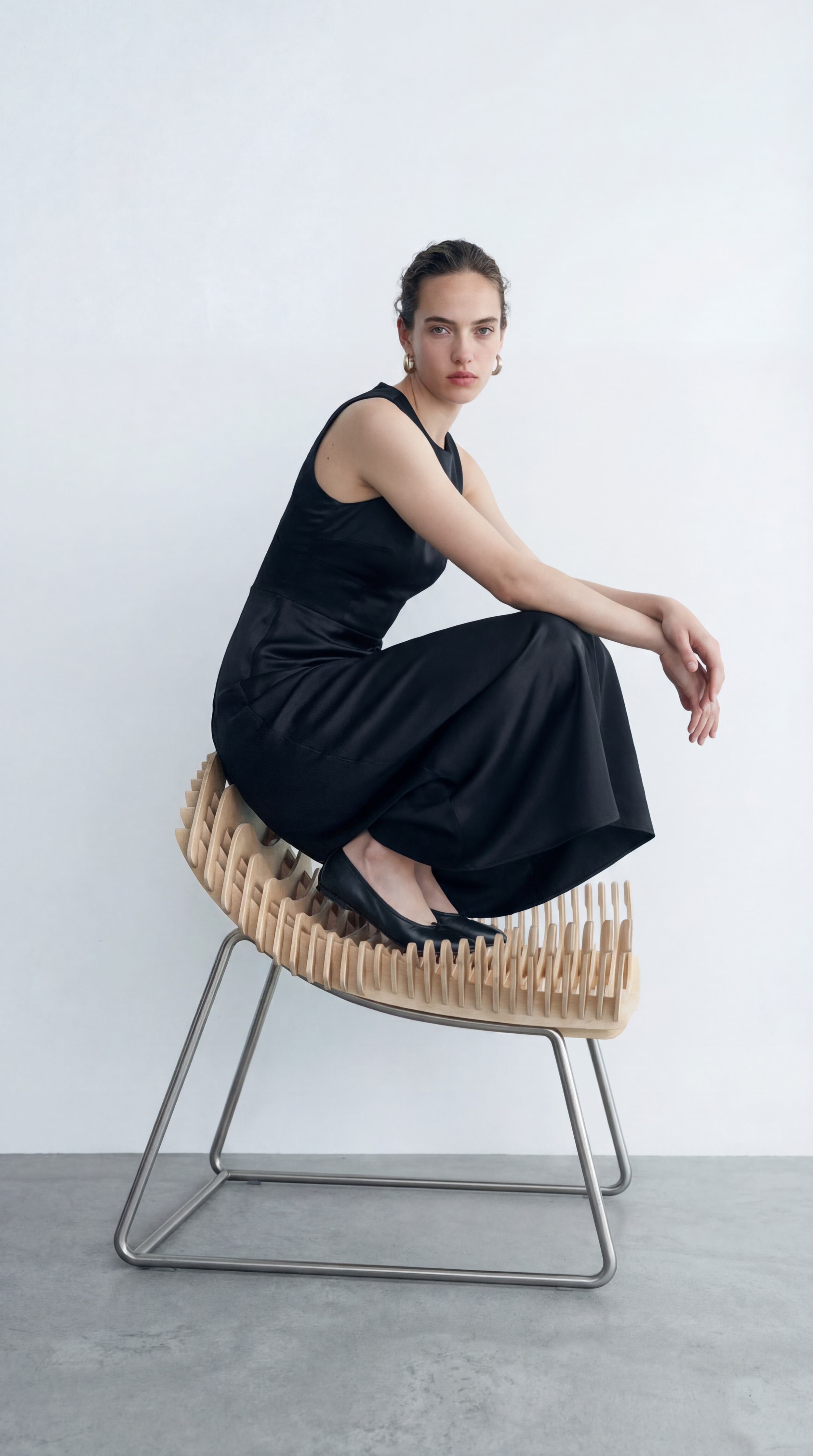 A woman with dark hair styled back, wearing a black sleeveless dress and black heels, sitting on a modern, curved wooden chair with metal legs against a plain white background.
