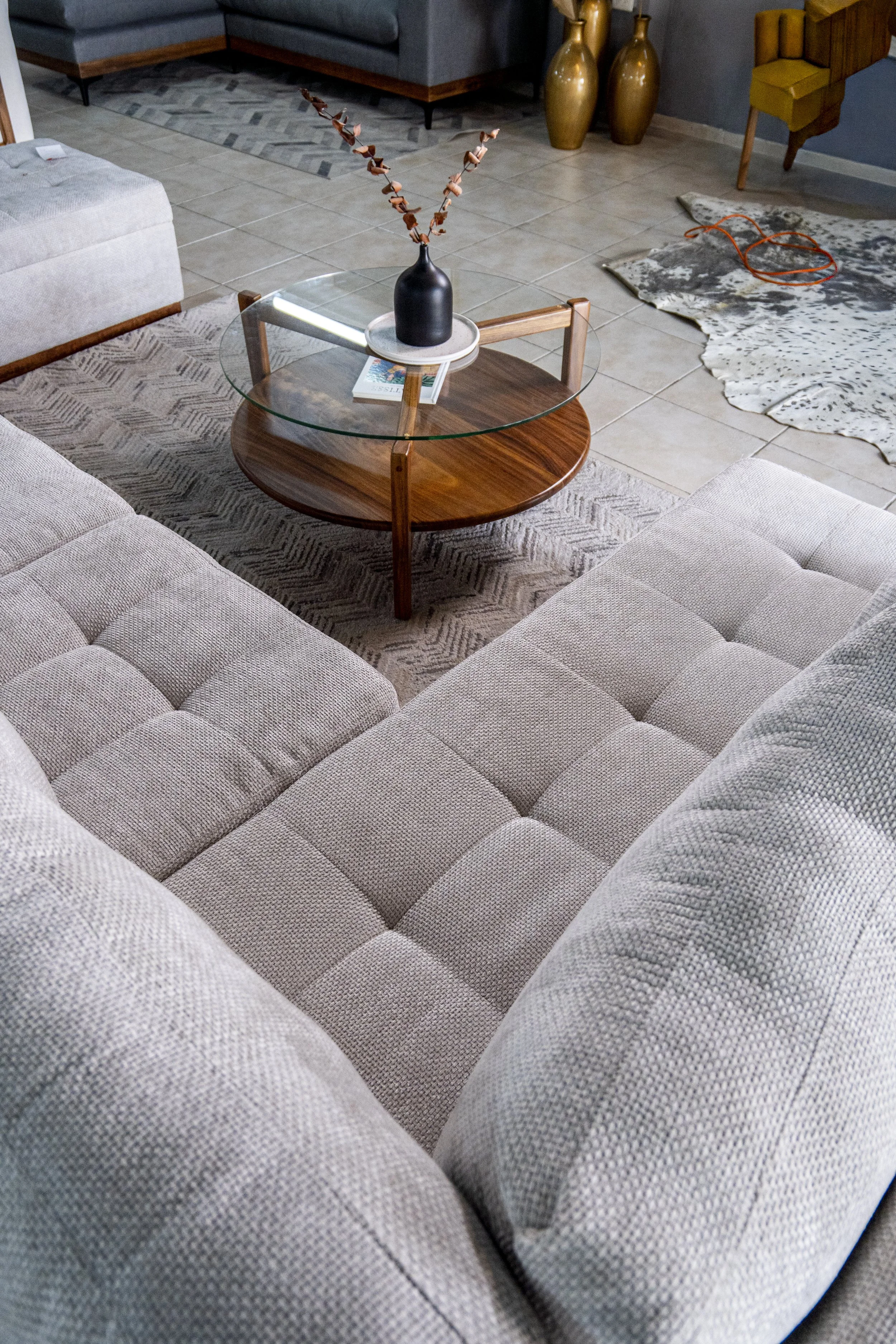Living room with beige upholstered sectional sofa, round glass coffee table, decorative vases, and area rug.