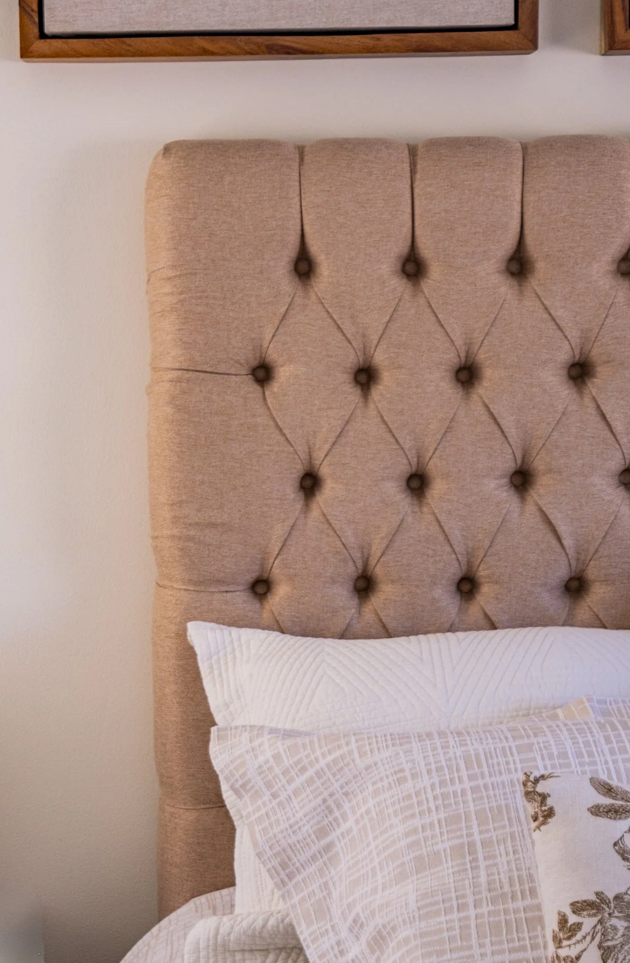 Close-up of a beige, tufted, upholstered headboard with diamond pattern, and a white or light-colored pillow with a textured pillowcase.