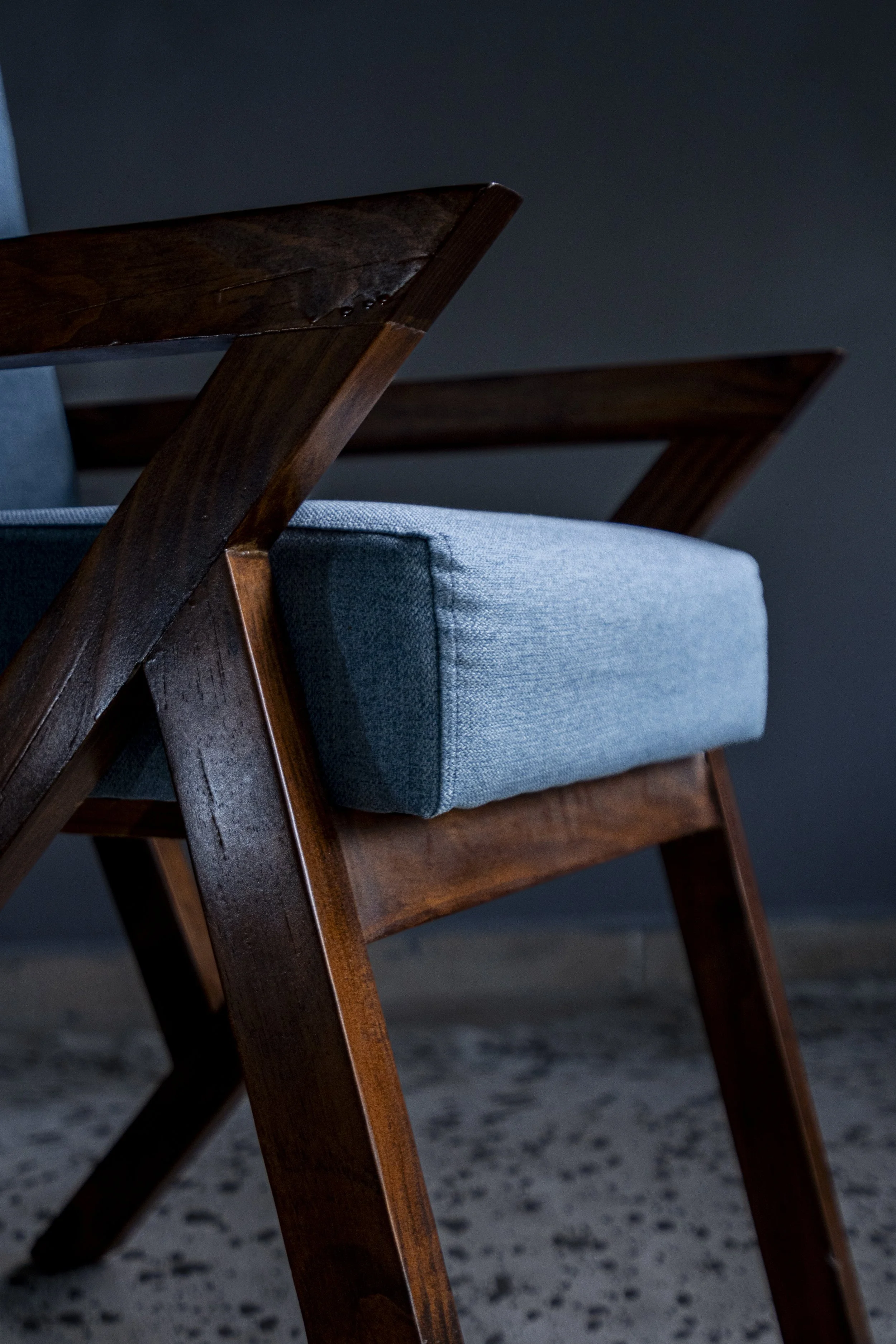 Close-up of a wooden chair with a blue fabric cushion on a dark floor with a dark background.