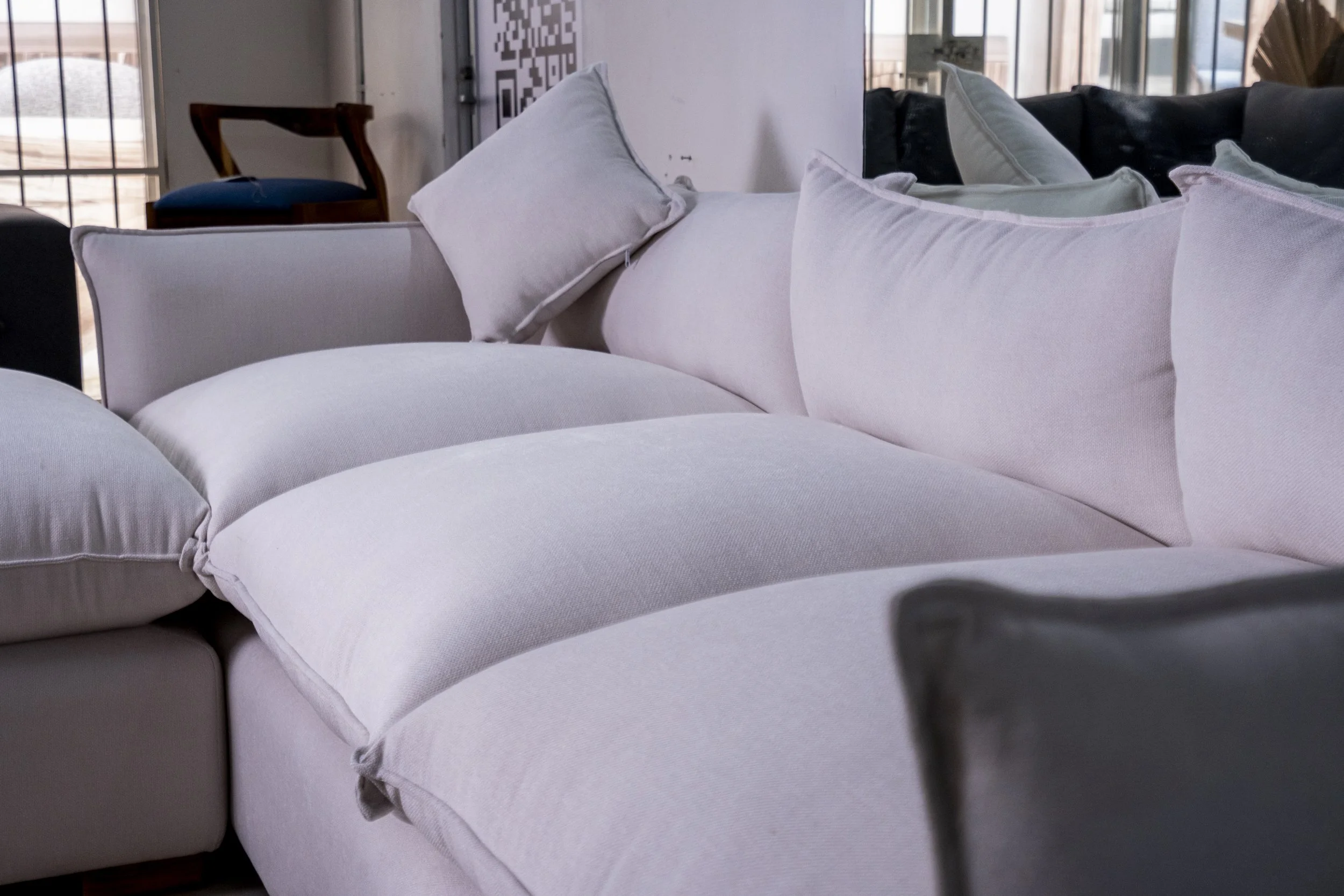 Close-up of a white fabric sofa with cushions in a room with large windows and wooden furniture.