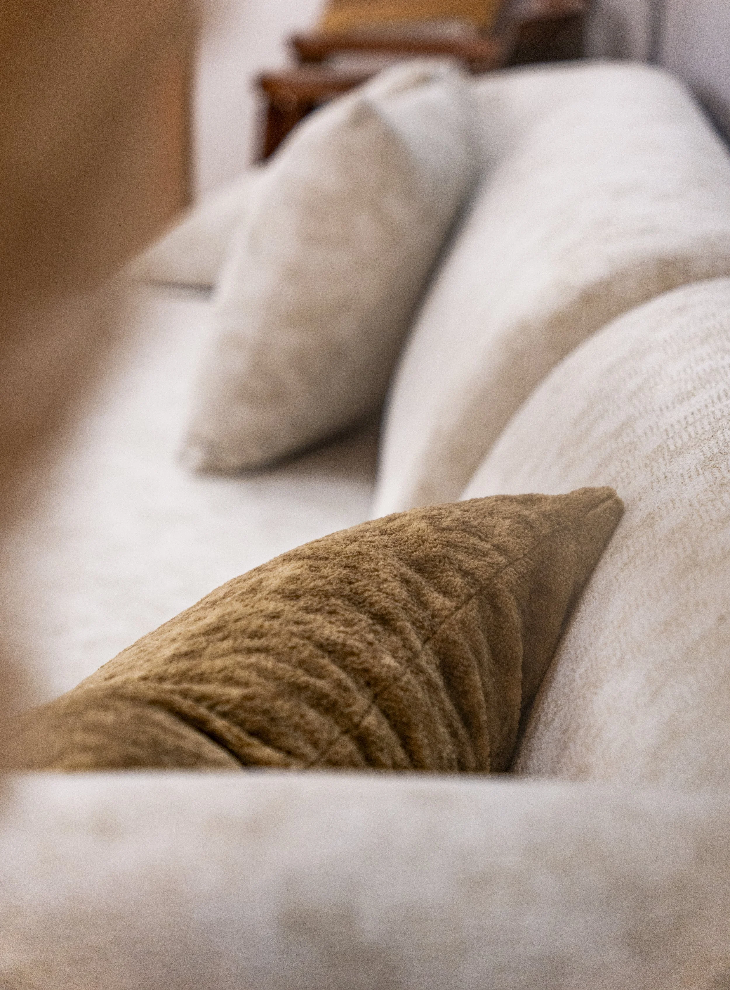 Close-up of a person's arm with a brown long sleeve resting on a light-colored sofa or bed with beige and white cushions.