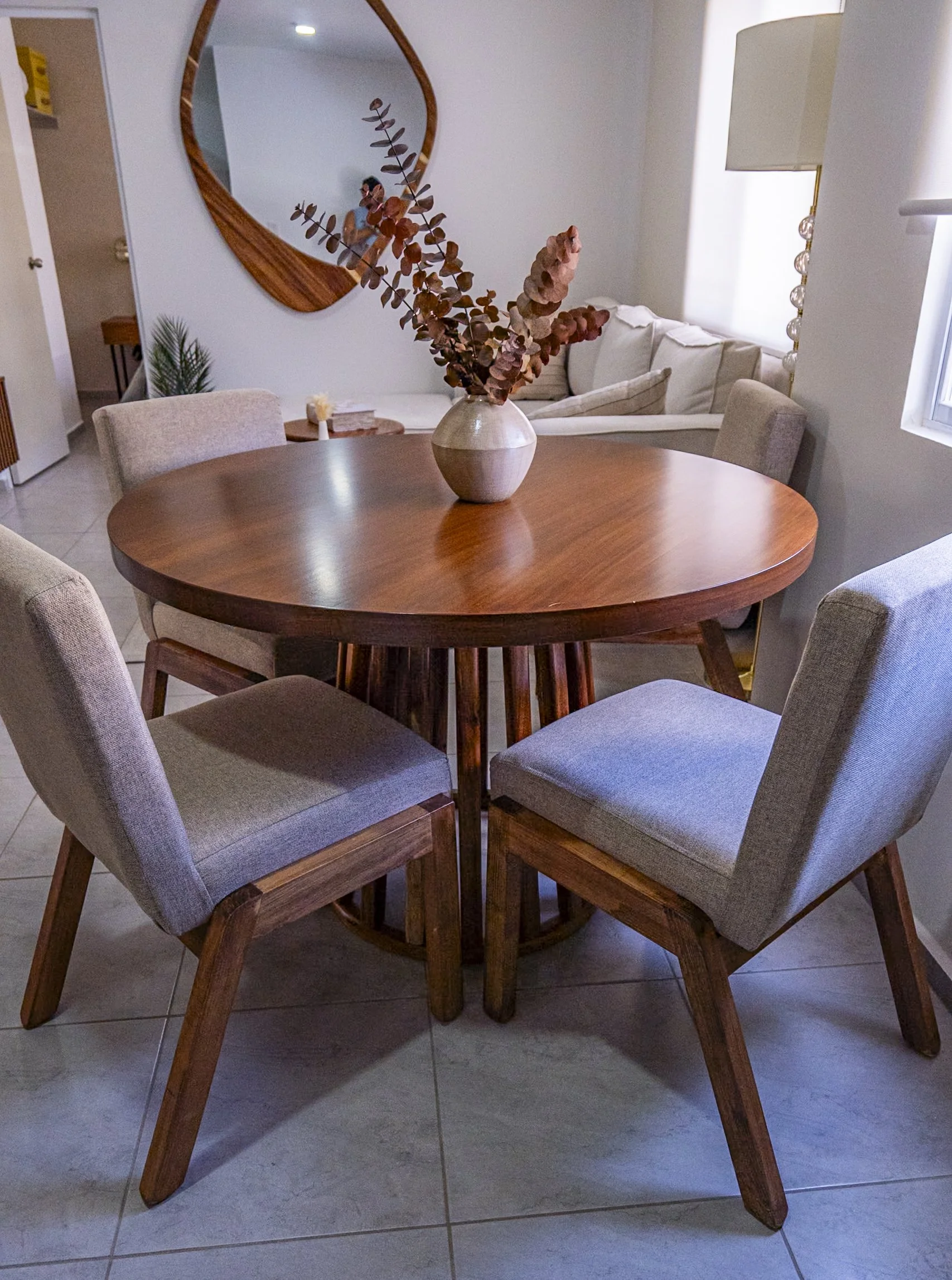 A round wooden dining table surrounded by six beige upholstered chairs in a room with tiled floor. A vase with dried reddish-brown eucalyptus branches is on the table. In the background, there is a white sofa, a tall lamp, and a large mirror on the wall.