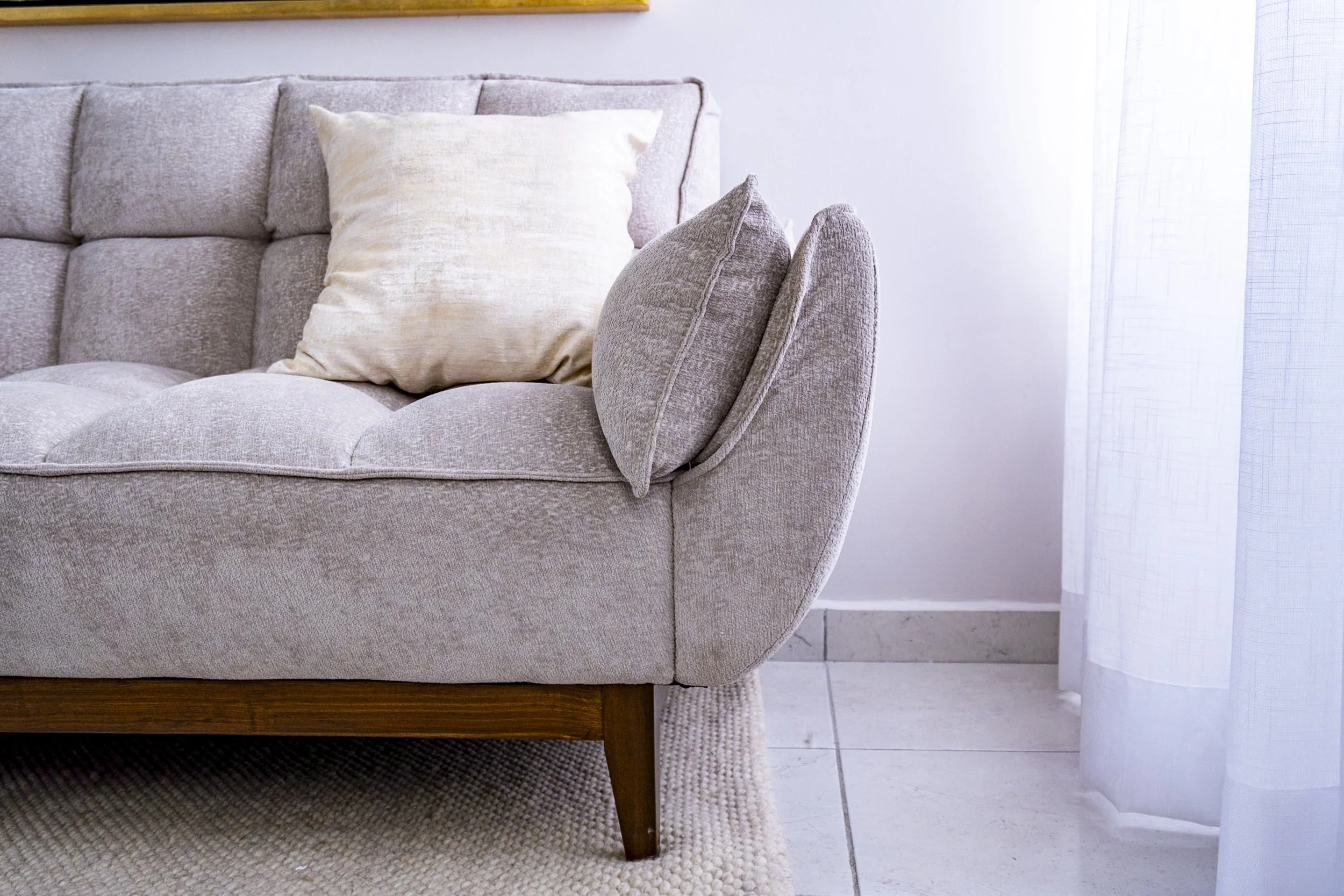 Close-up of a beige fabric sofa with two cushions, one plain and one with textured pattern, in front of a white wall with a yellow border at the top and a white curtain on the right side.