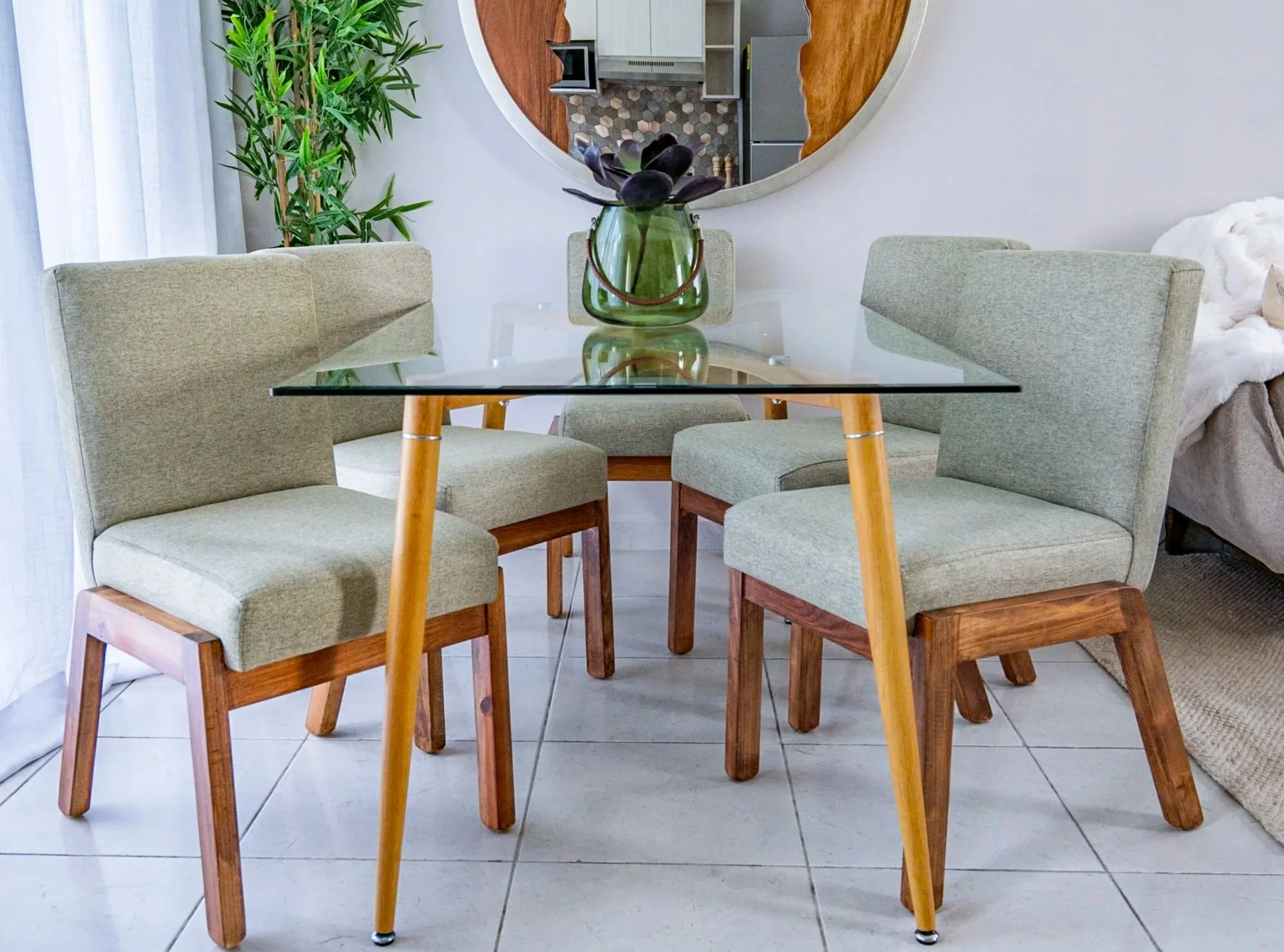 A dining room with a glass table, four green upholstered chairs with wooden legs, a large mirror on the wall, a green vase with purple succulents, a potted plant, and a view into a kitchen and living area.