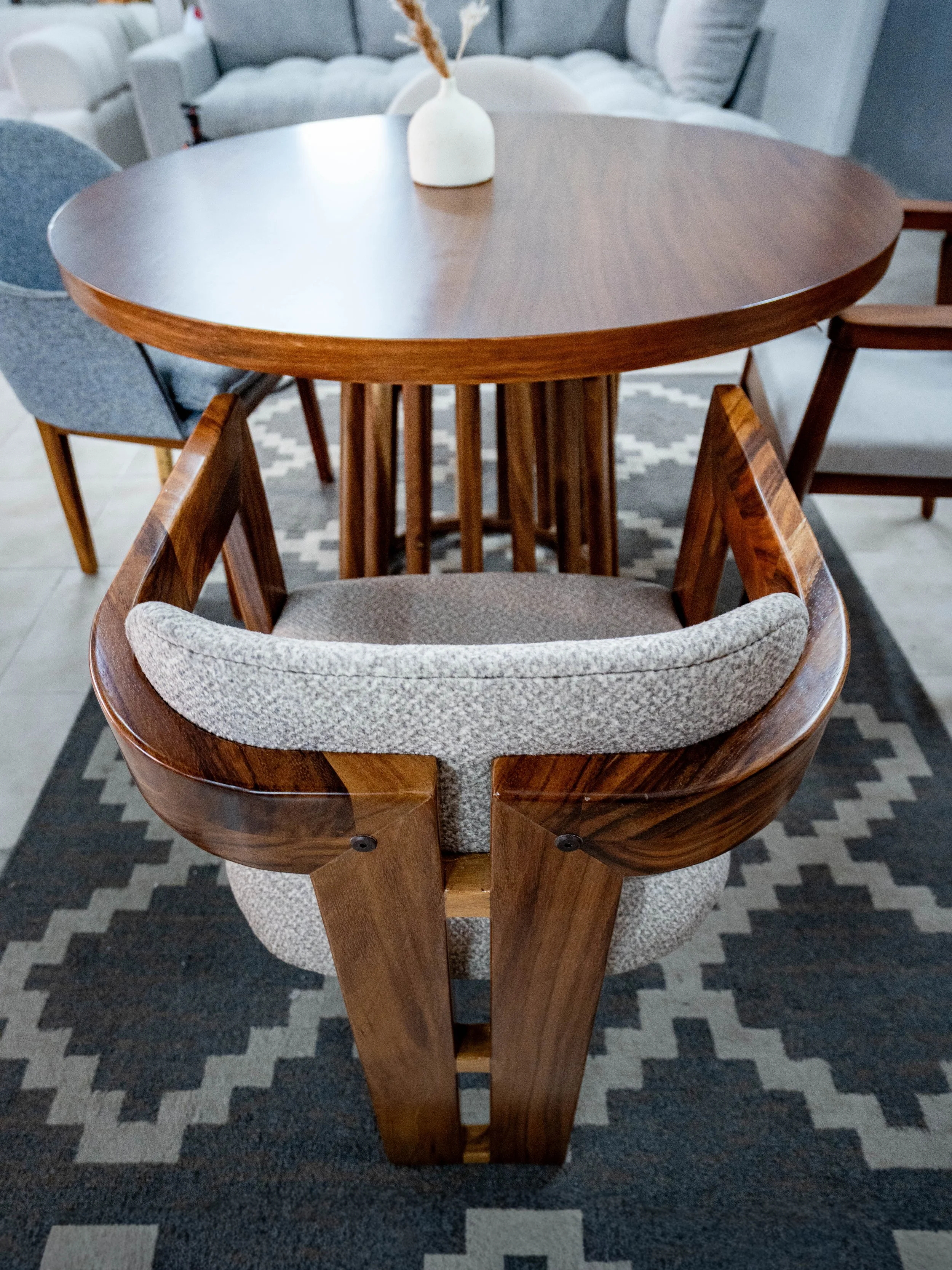 A wooden dining table with a white vase containing dried decorative grasses, surrounded by upholstered chairs with wooden arms, on a black and white geometric rug.