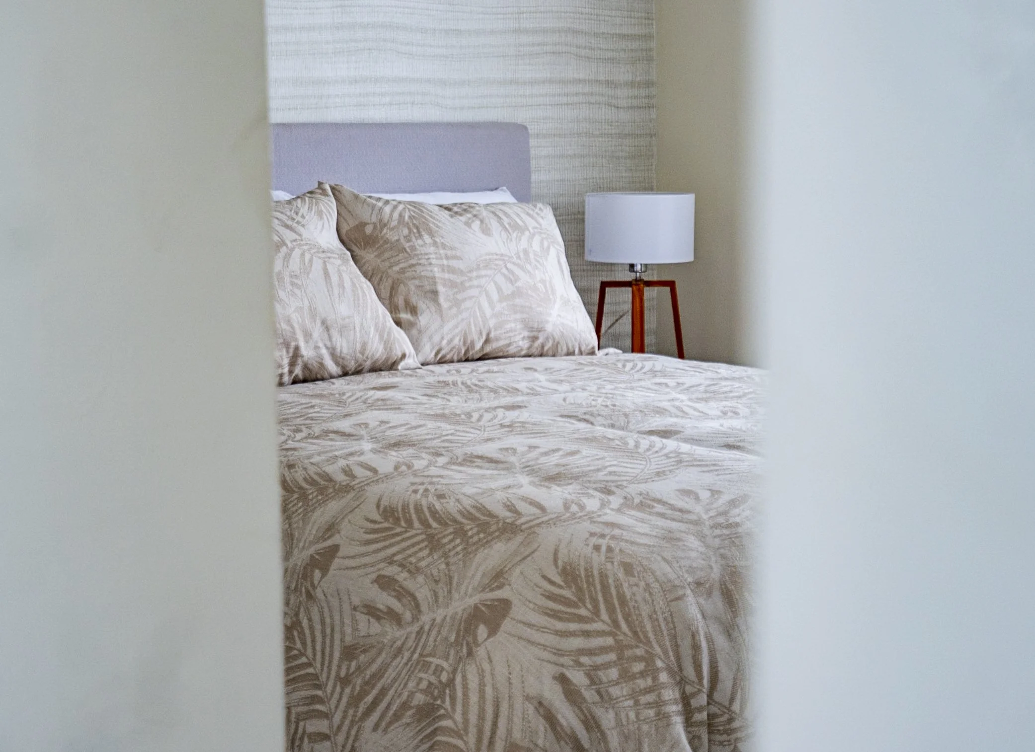 Part of a bedroom with a bed that has beige and white patterned bedding, a matching pillow, a gray headboard, a bedside table with a white lamp, and a wallpapered accent wall.