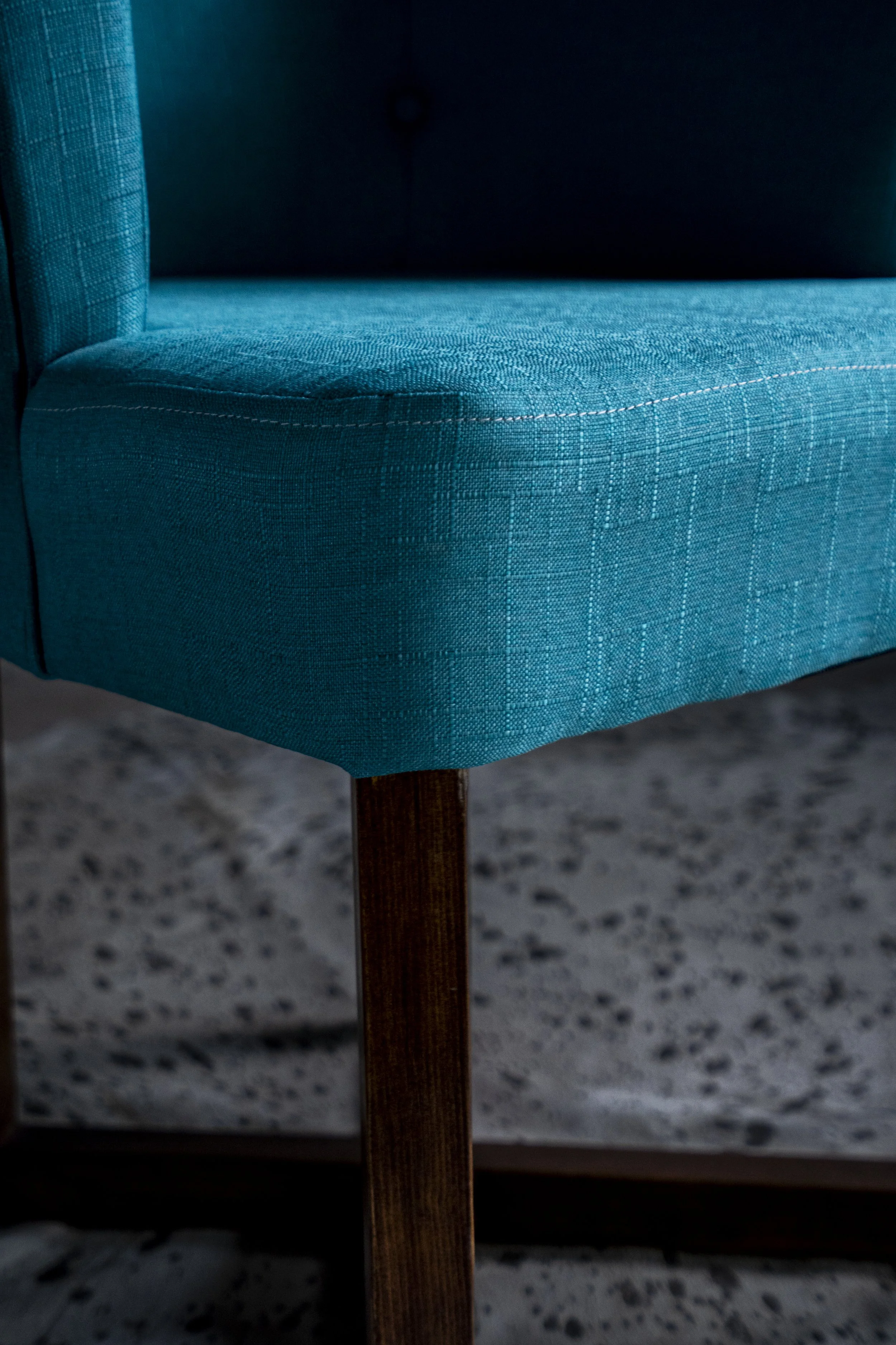 Close-up of a teal upholstered chair with wooden legs on a gray speckled floor.