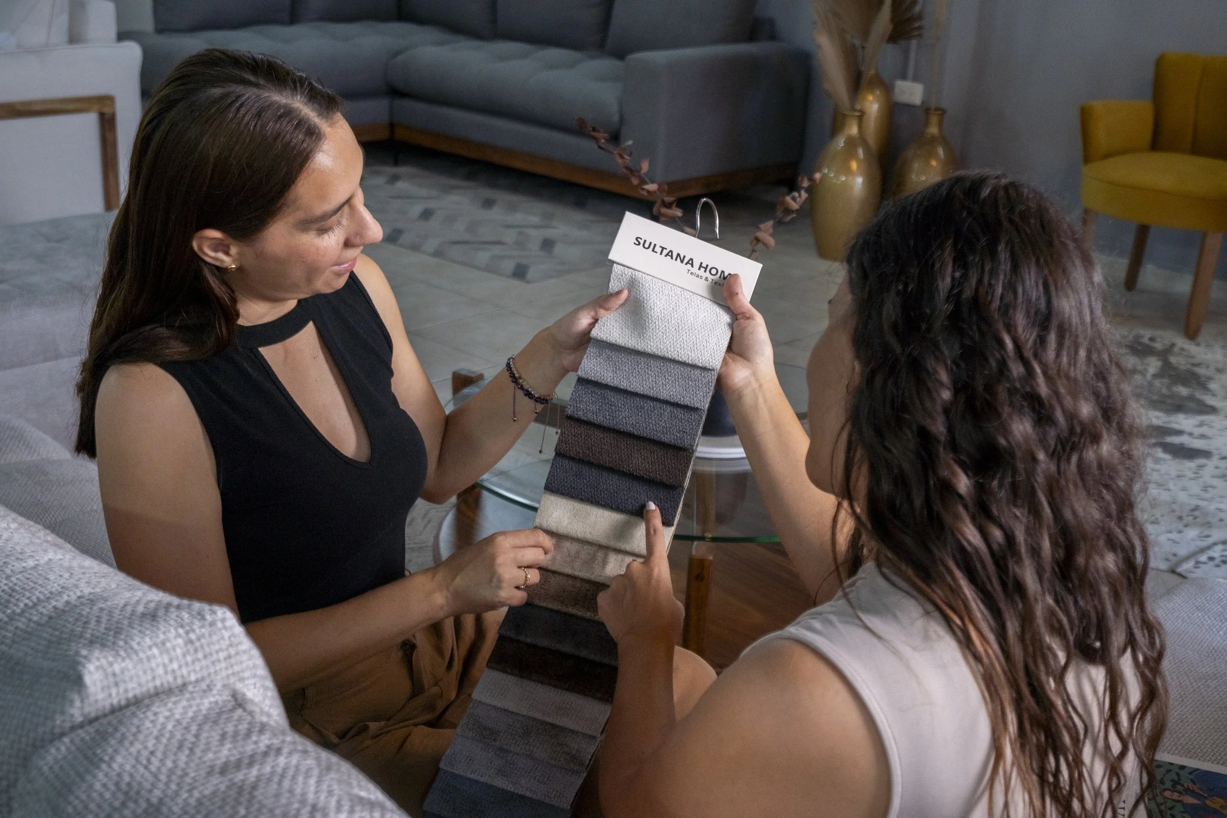 Two women sitting on a couch in a living room, holding a fabric sample book and looking at it together. One woman has brown hair with highlights, wearing a white top, and the other has brown, wavy hair, wearing a black sleeveless top.