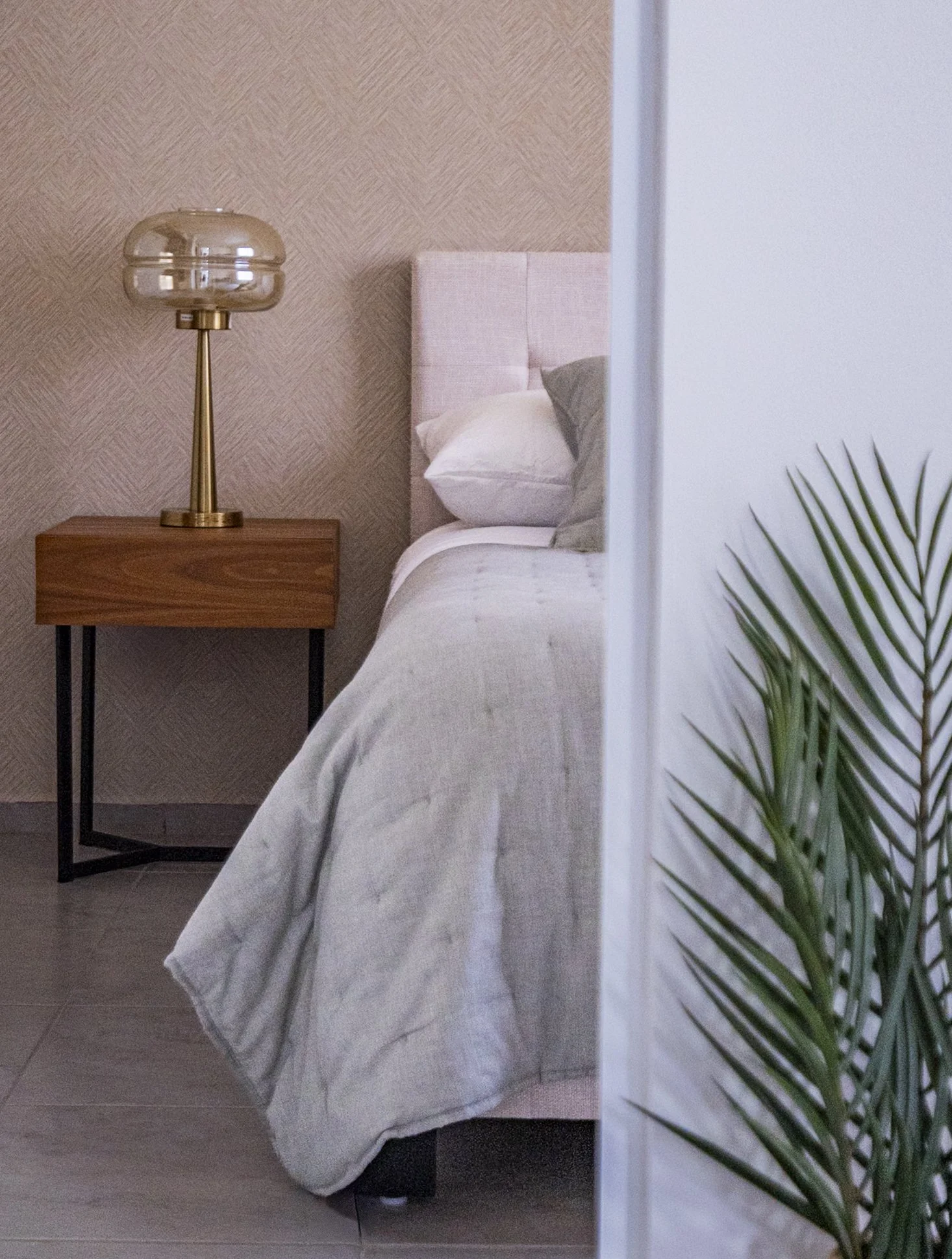 A glimpse of a bedroom with a wooden nightstand, a modern table lamp with a glass top, a pink upholstered headboard, white bedsheets, and a gray pillow. A green potted plant is visible in the foreground.
