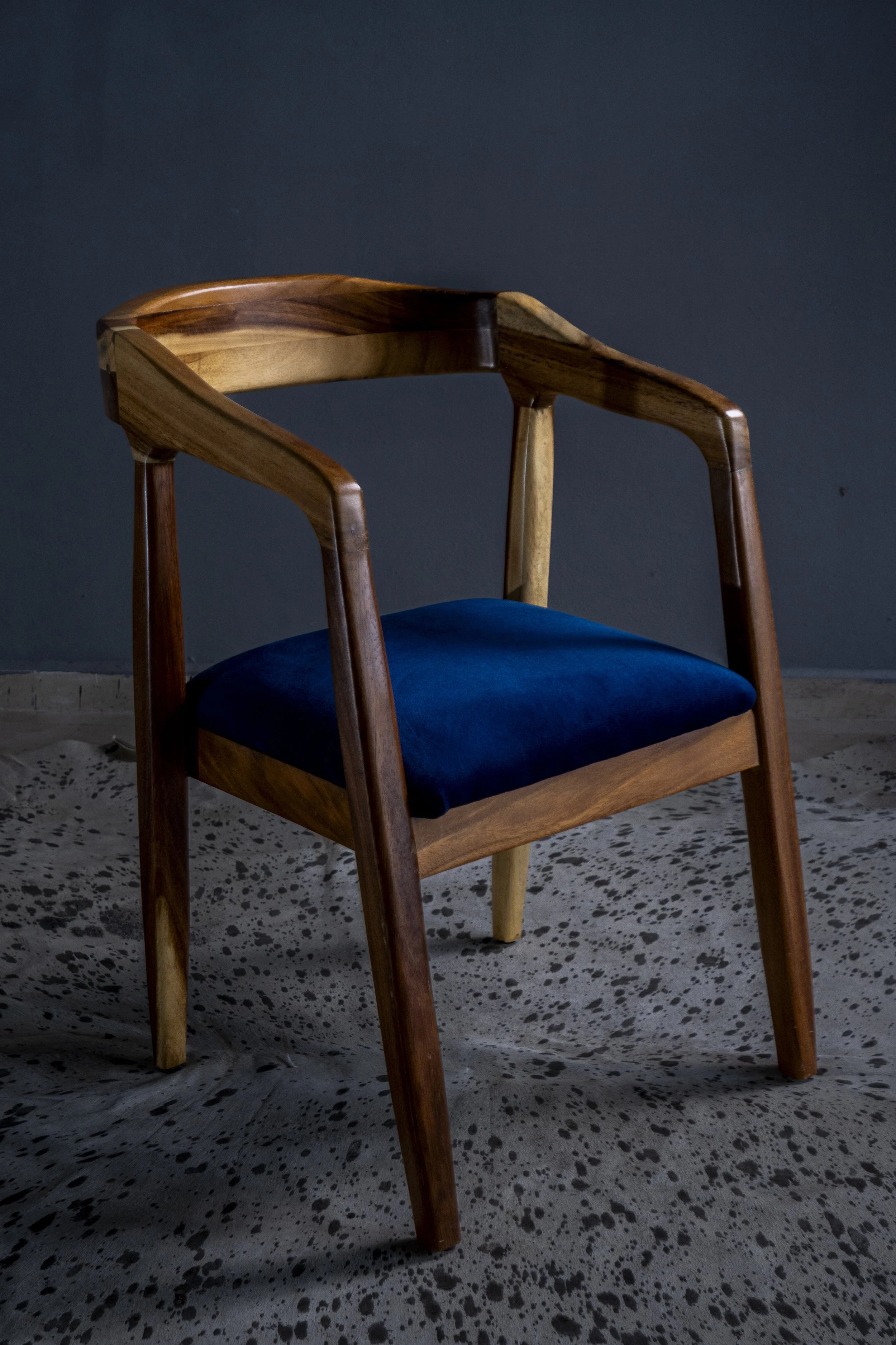 A wooden chair with a dark blue cushioned seat positioned on a white and black speckled floor against a dark wall.