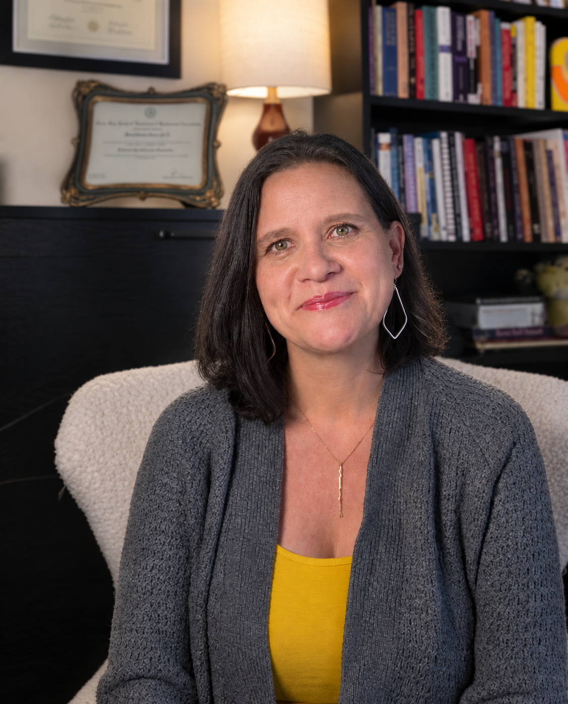 A woman with shoulder-length dark hair, wearing a gray cardigan over a yellow top, sitting on a cream-colored chair in a room with bookshelves and a framed certificate on the wall behind her.