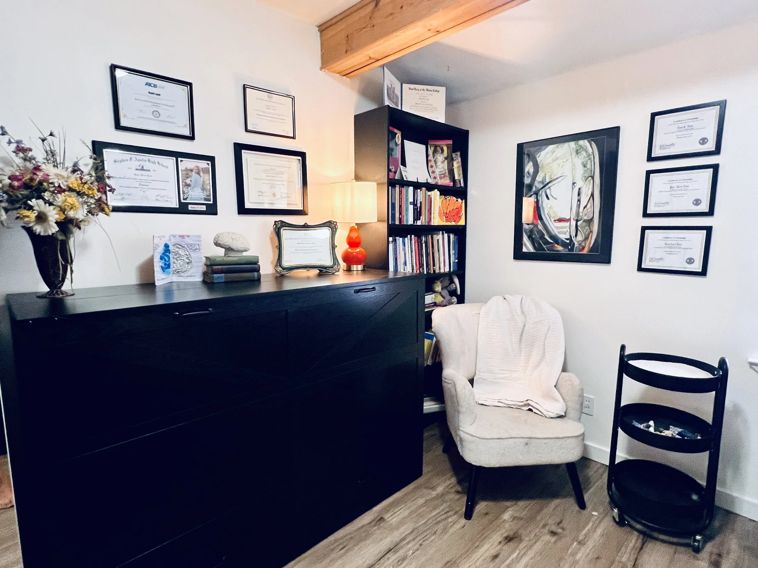 A corner of a room with a white wall decorated with framed certificates and a colorful abstract painting. There is a black dresser with a vase of dried flowers, a lamp, and some books. A white cushioned armchair is beside the dresser, and a black wooden chair is nearby. The room has a wooden floor.