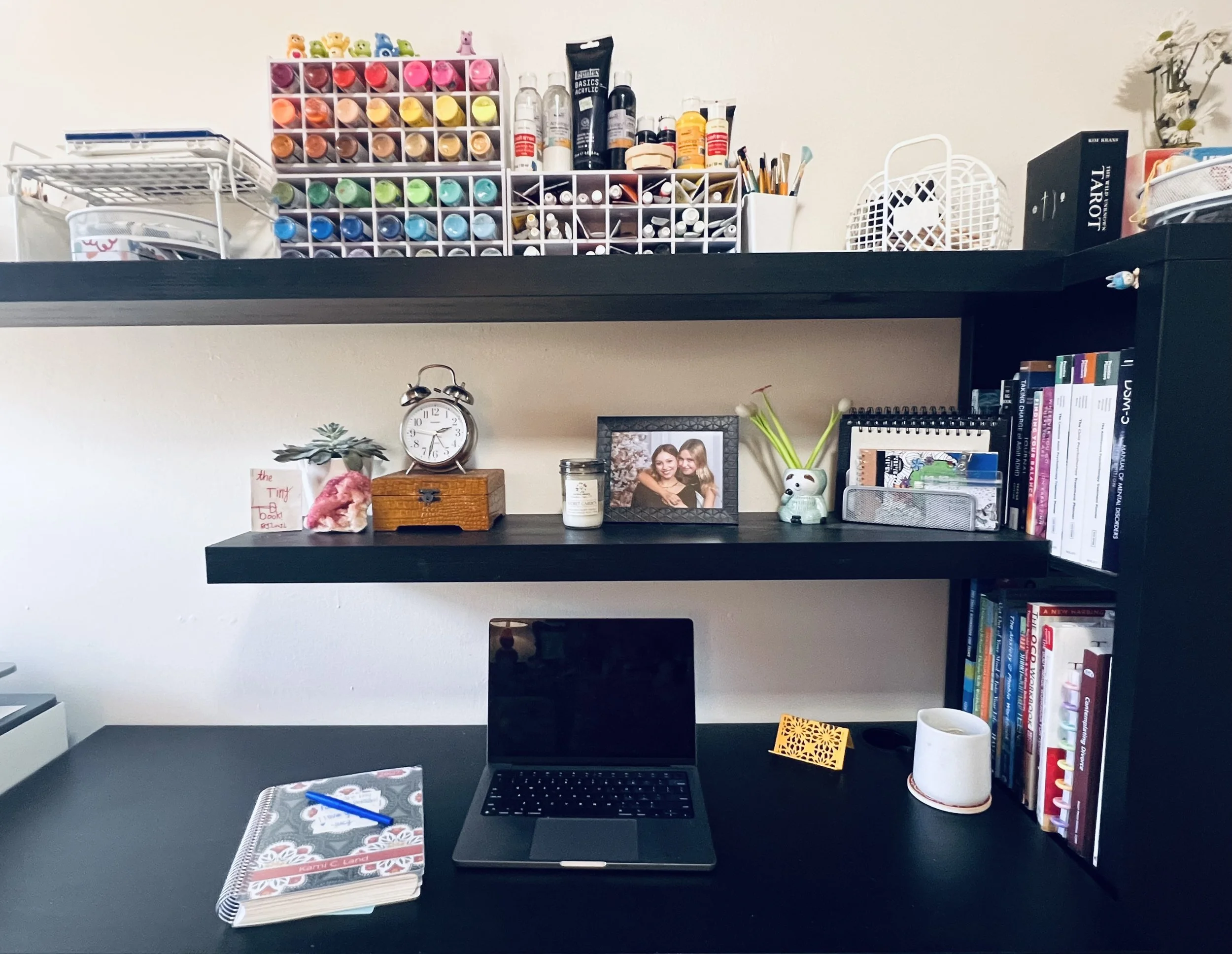 A computer desk with a notebook, pen, keyboard, mouse pad, and a digital drawing tablet. Above the desk, a black shelf holds decorative plants, a clock, framed photos, books, and office supplies. Shelves contain paint bottles, art supplies, and books.