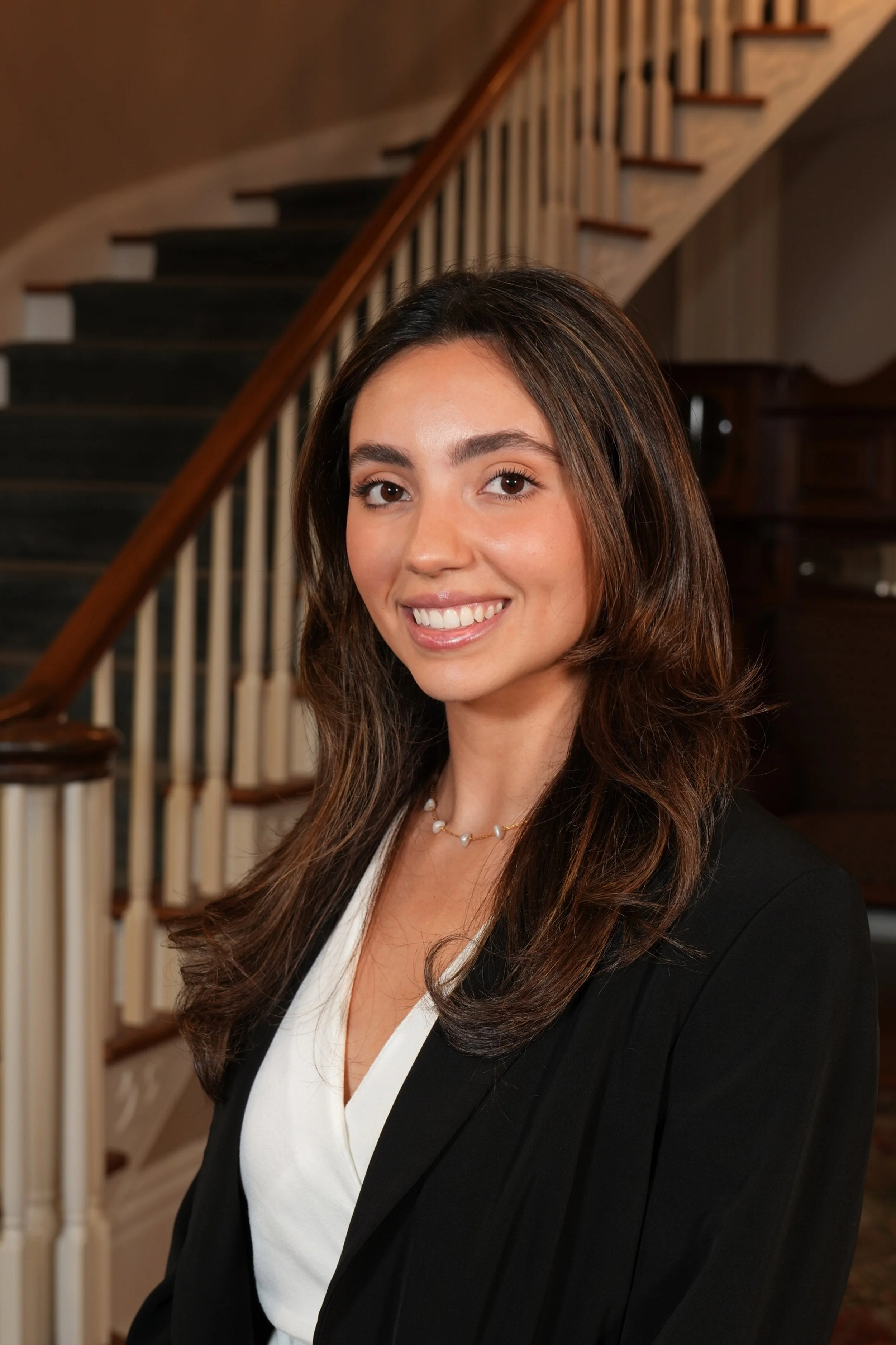 A woman with long brown hair and brown eyes smiling, wearing a black blazer with white blouse, standing indoors near a staircase with wooden handrail.