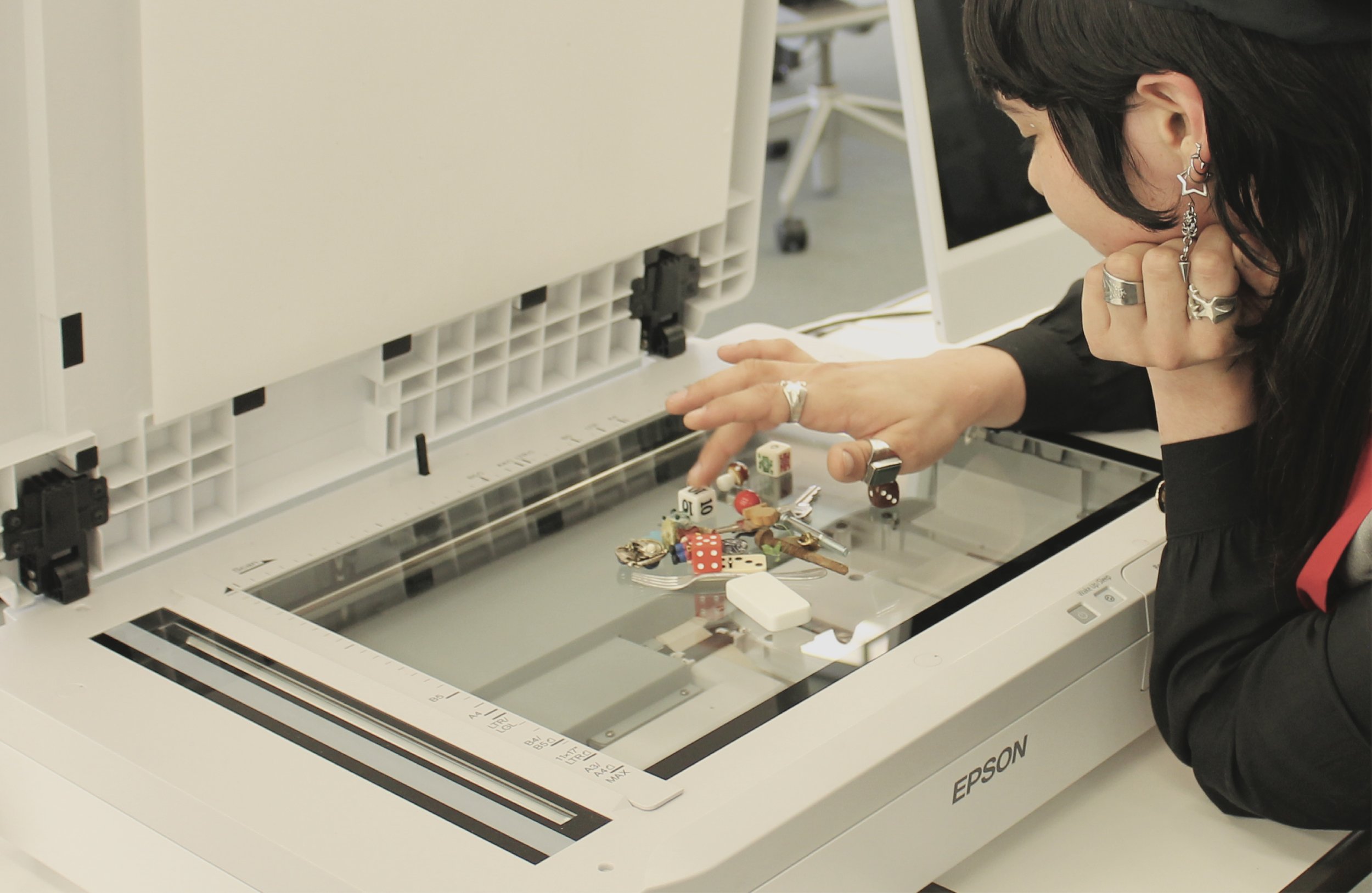 A person with black hair and multiple earrings is leaning over an Epson scanner, examining a collection of small objects including dice, figurines, and other items on the scanner bed.
