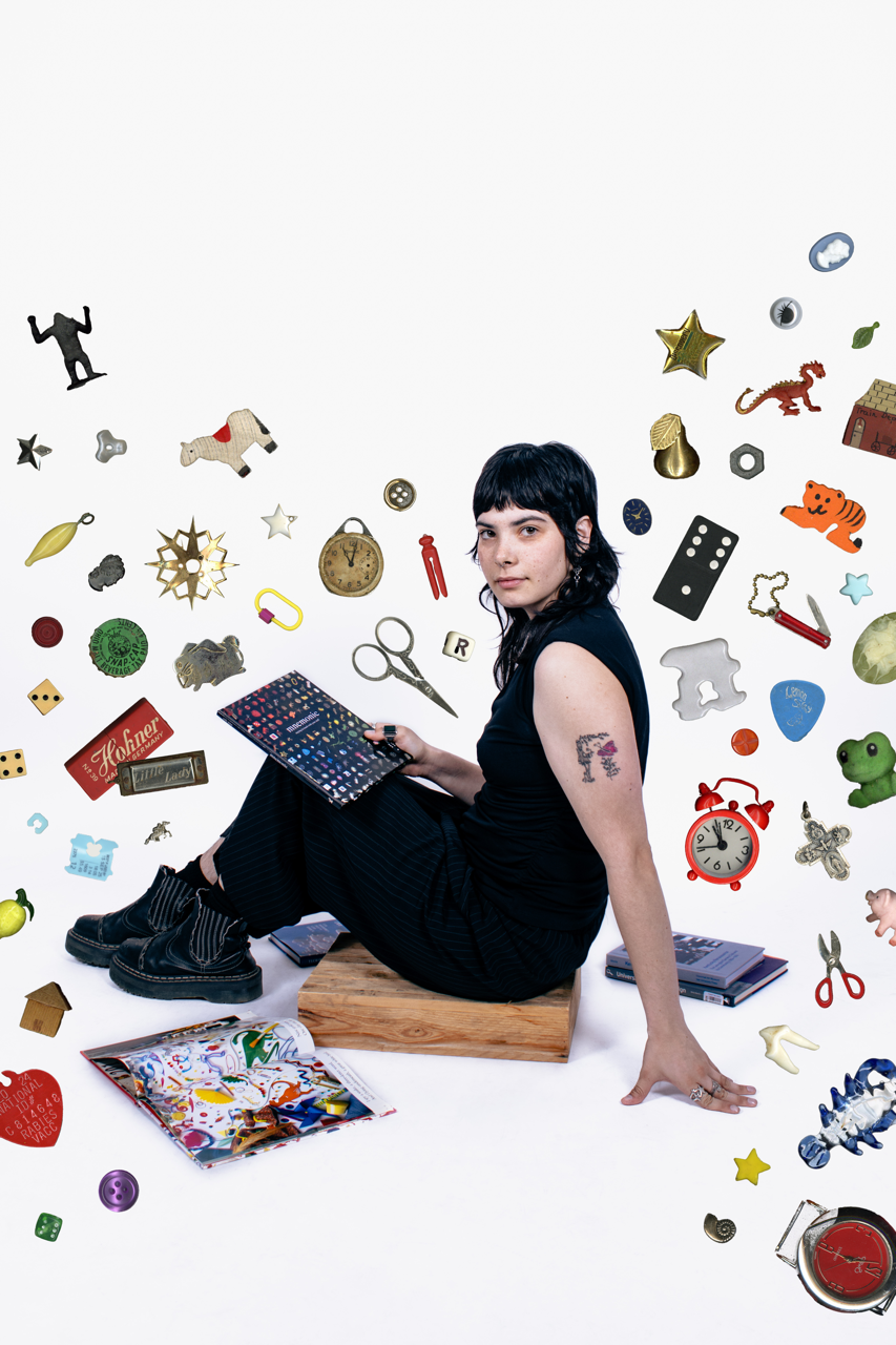 Young woman sitting on a wooden block surrounded by scattered small objects and toys, holding a tablet, with a plain white background.