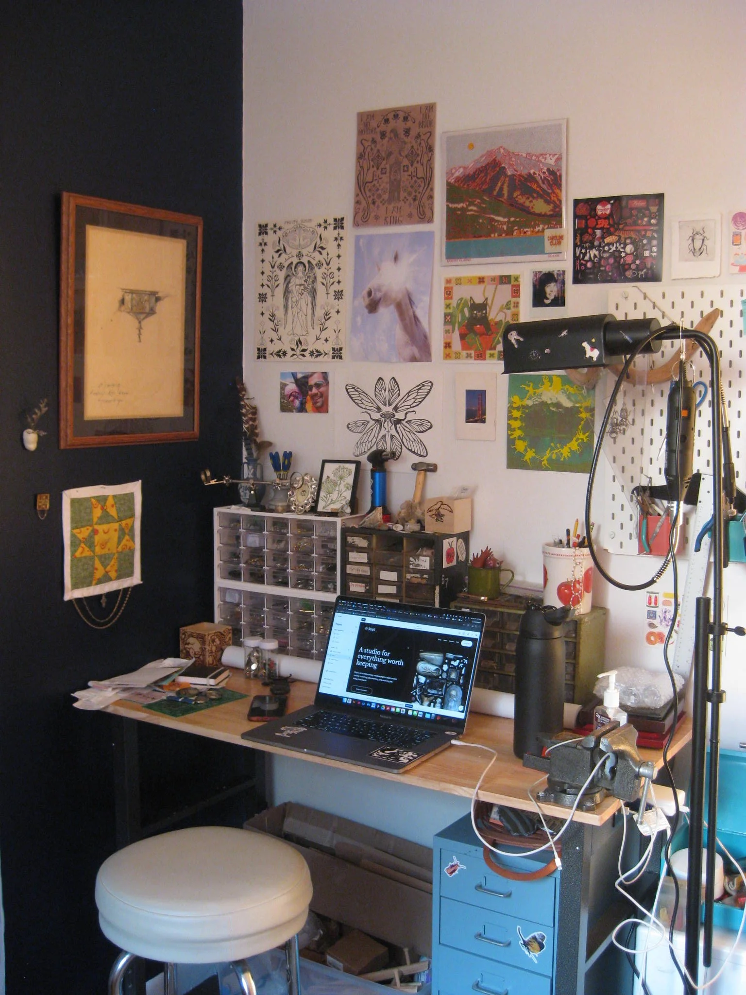 A workspace with a wooden desk holding a laptop open, a black lamp, and various supplies. A small blue filing cabinet with stickers, and a white stool with a round seat are nearby. The background wall displays multiple pieces of art, photos, and decorations, including a large landscape painting, a llama picture, and other artwork. A pegboard with tools and organized items is on the right side.