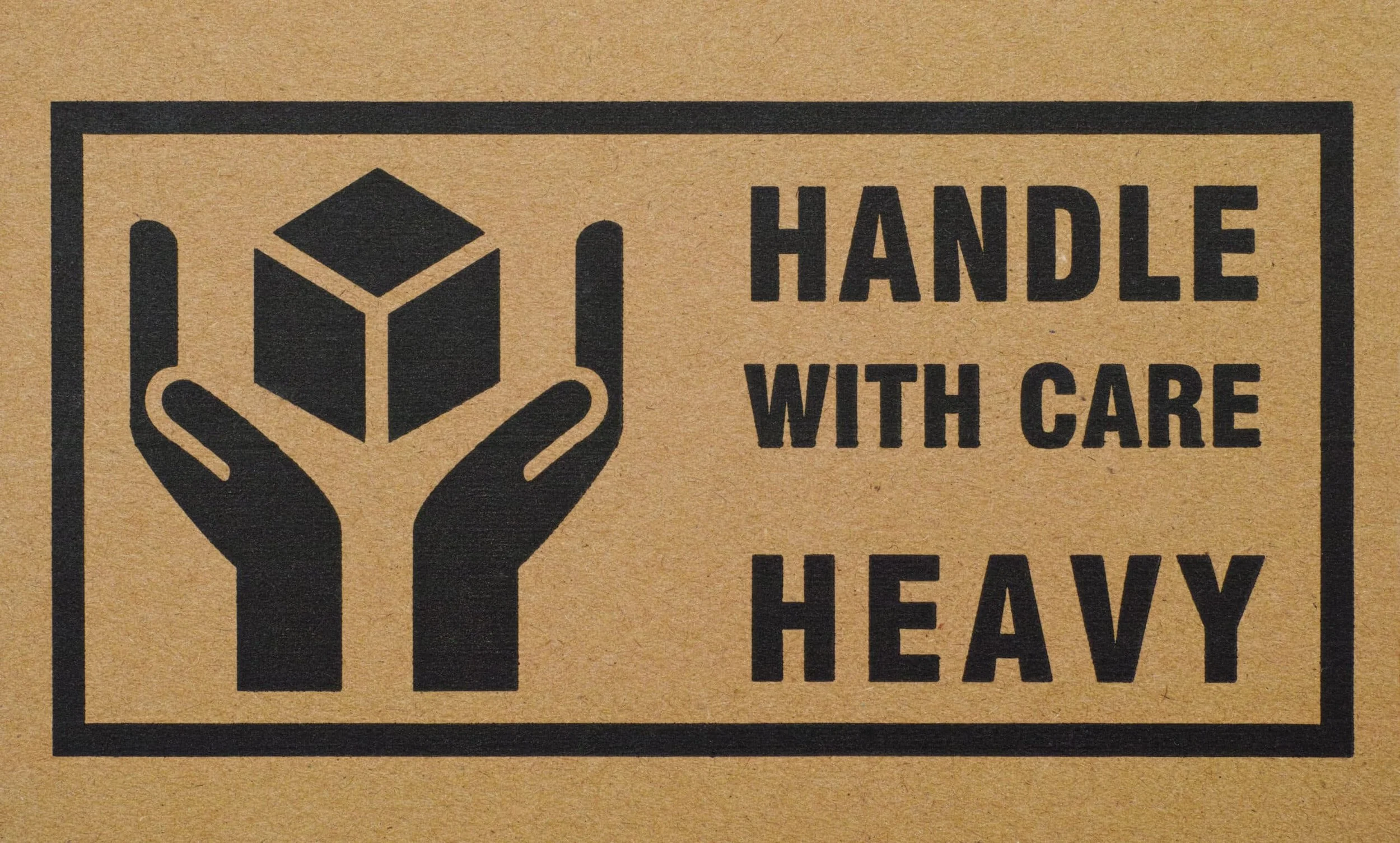 A cardboard box with a black printed graphic of a hand holding a cube and the text 'HANDLE WITH CARE HEAVY'.