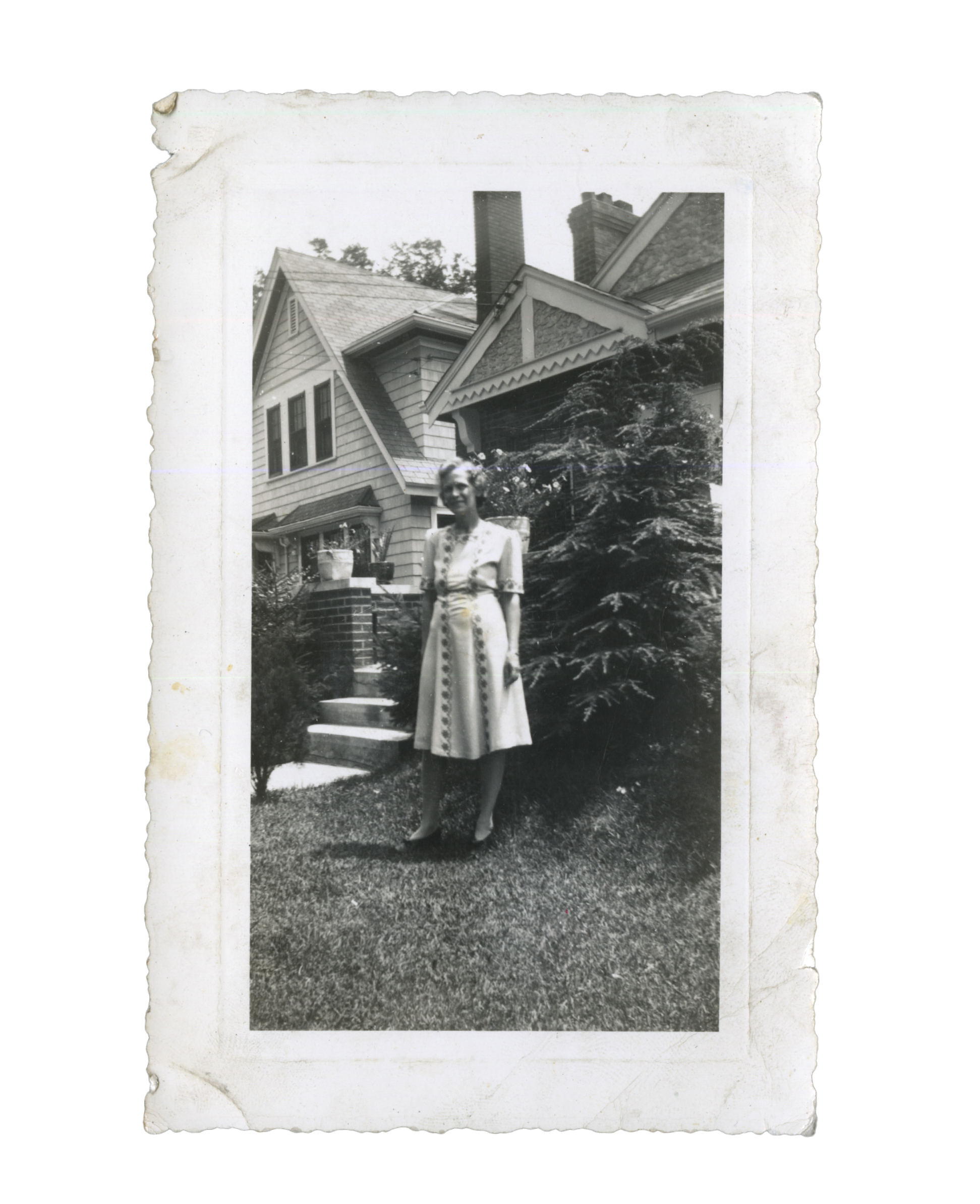 Black and white photo of a woman standing outdoors in front of a house with multiple gables and chimneys, trees, and a garden.
