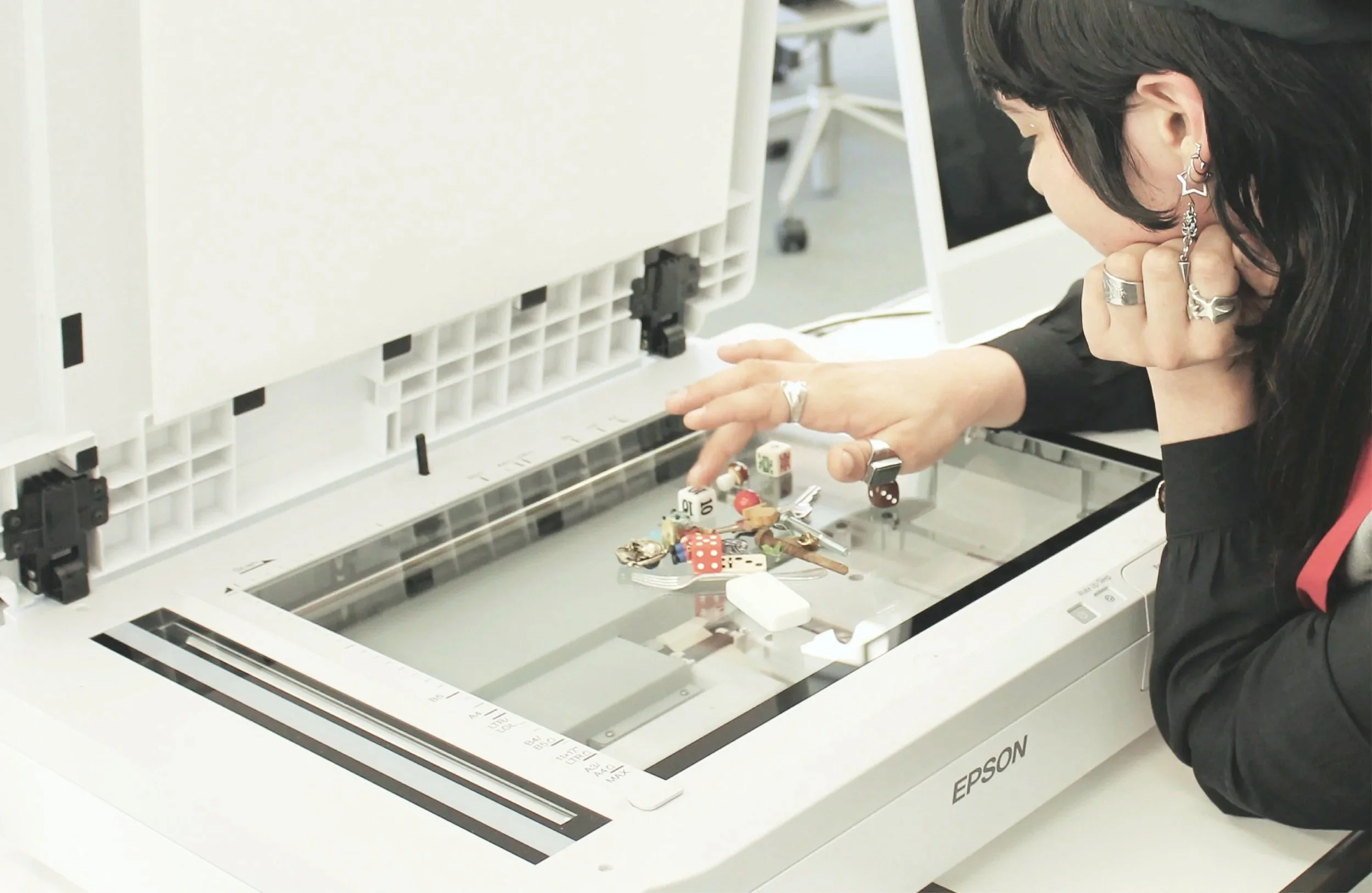 A woman with dark hair and multiple rings is leaning over an Epson scanner, looking at objects on the scanner bed, including small figurines and game pieces.