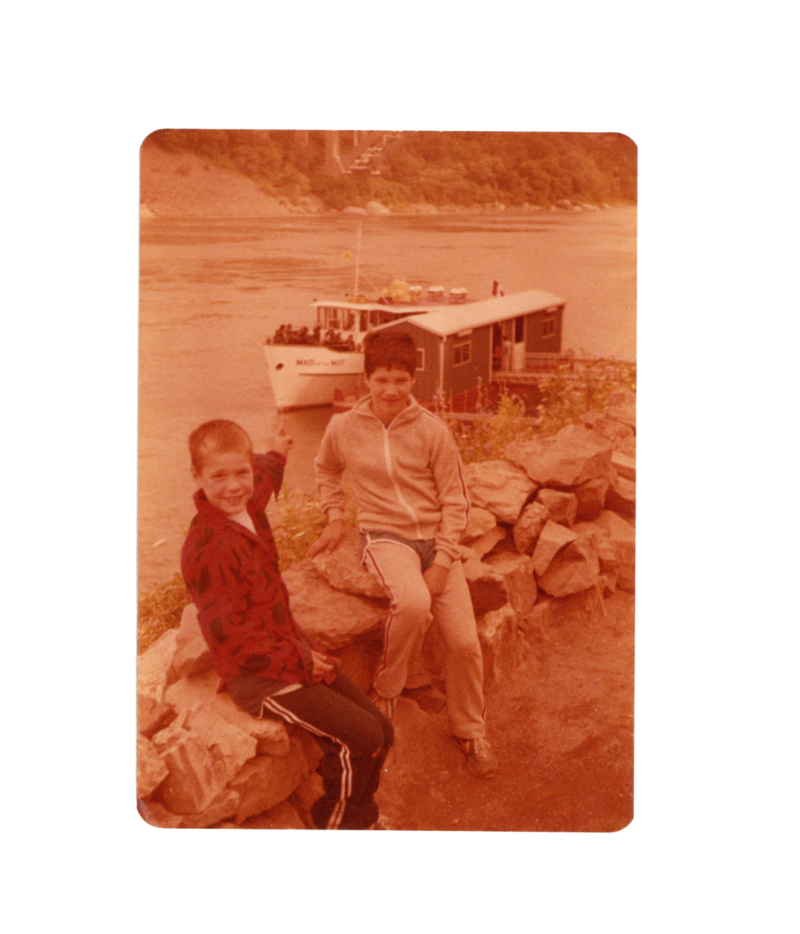 Two boys sitting on rocks beside a river, with a boat docked behind them and a houseboat in the water.