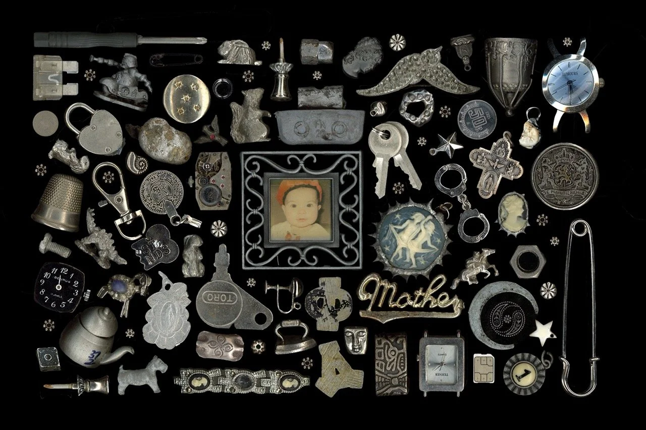 A collection of assorted small objects including jewelry, watches, a toy horse, a photo of a baby in a decorative frame, keys, spools, and various metal trinkets arranged on a black background.