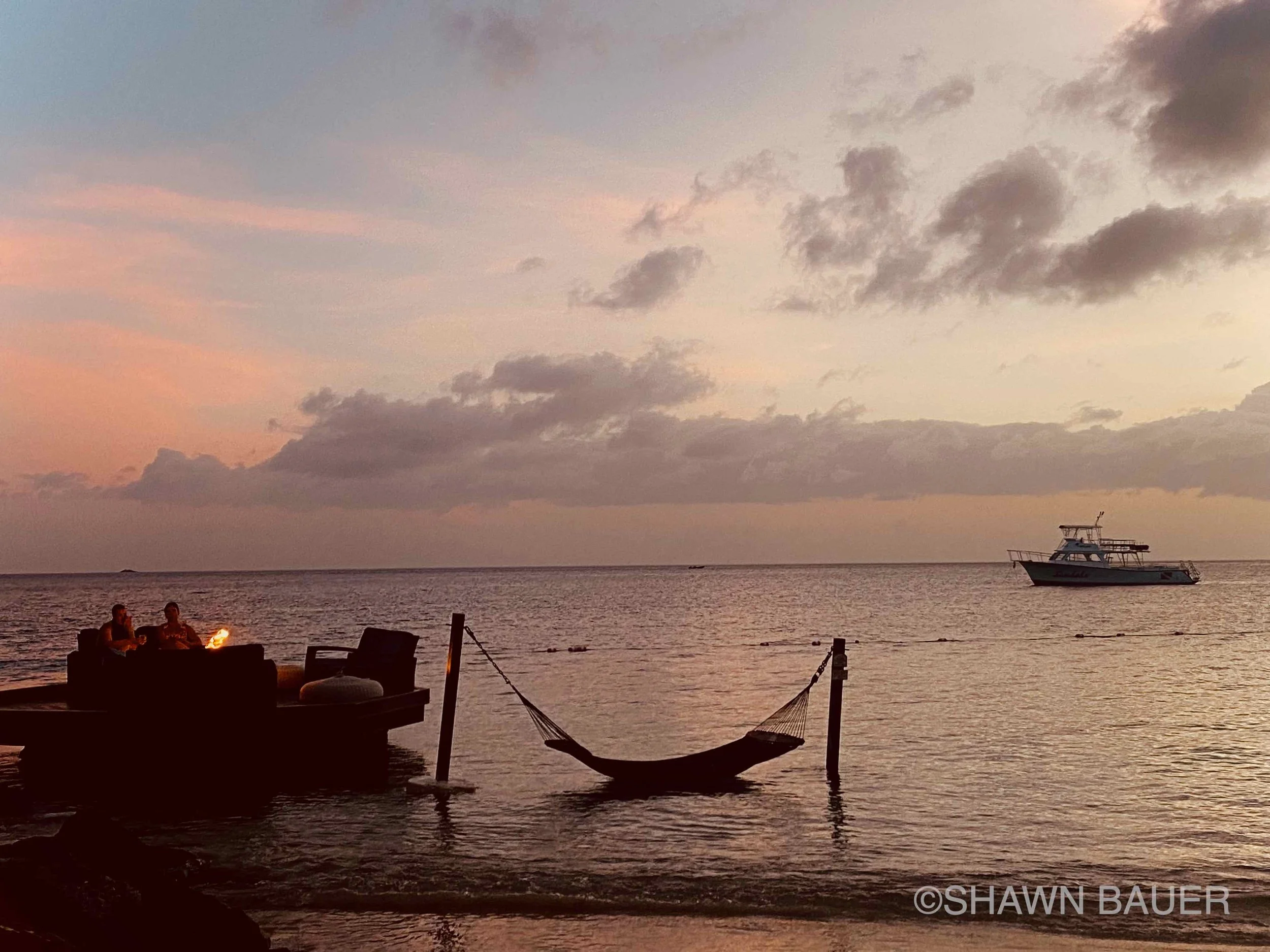 A tranquil scene at sunset with a boat on the water, two people sitting by a fire on the shore, and a hammock hanging between two posts near the water.