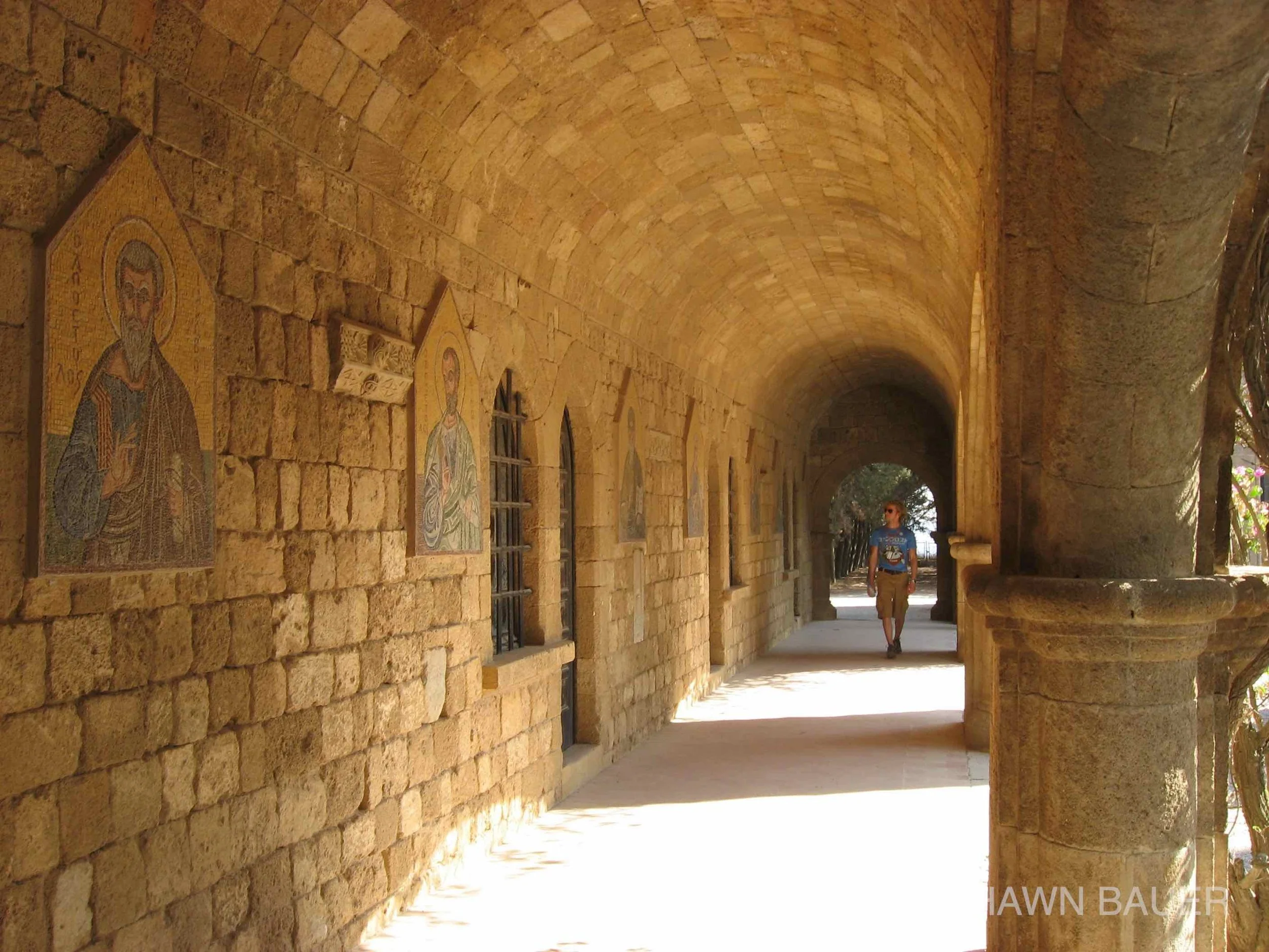 A stone archway with religious mosaic images on the wall, a walkway, and a person walking underneath.