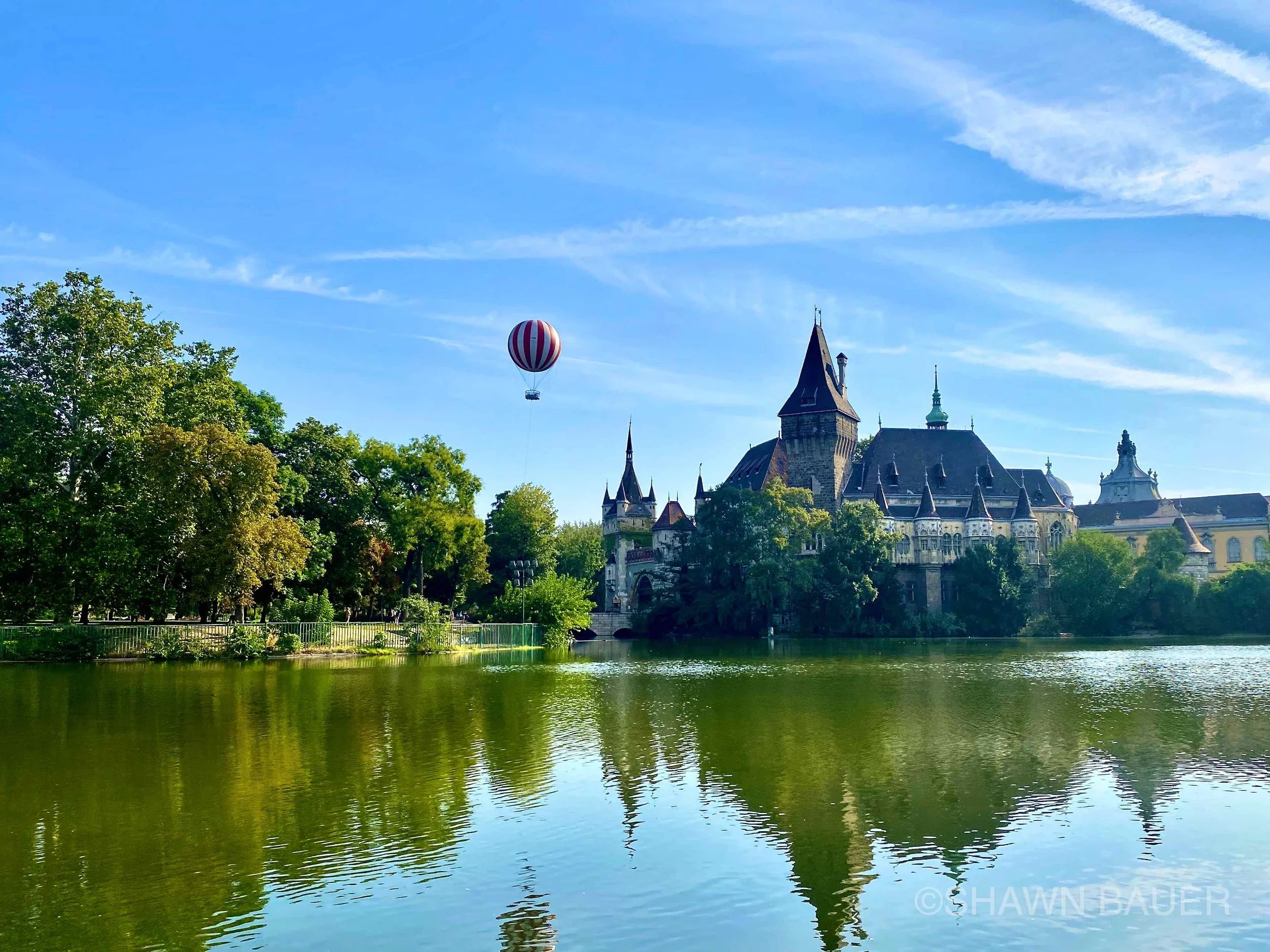 A hot air balloon with red and white stripes flying over a castle on a sunny day, with green trees and a river in the foreground.