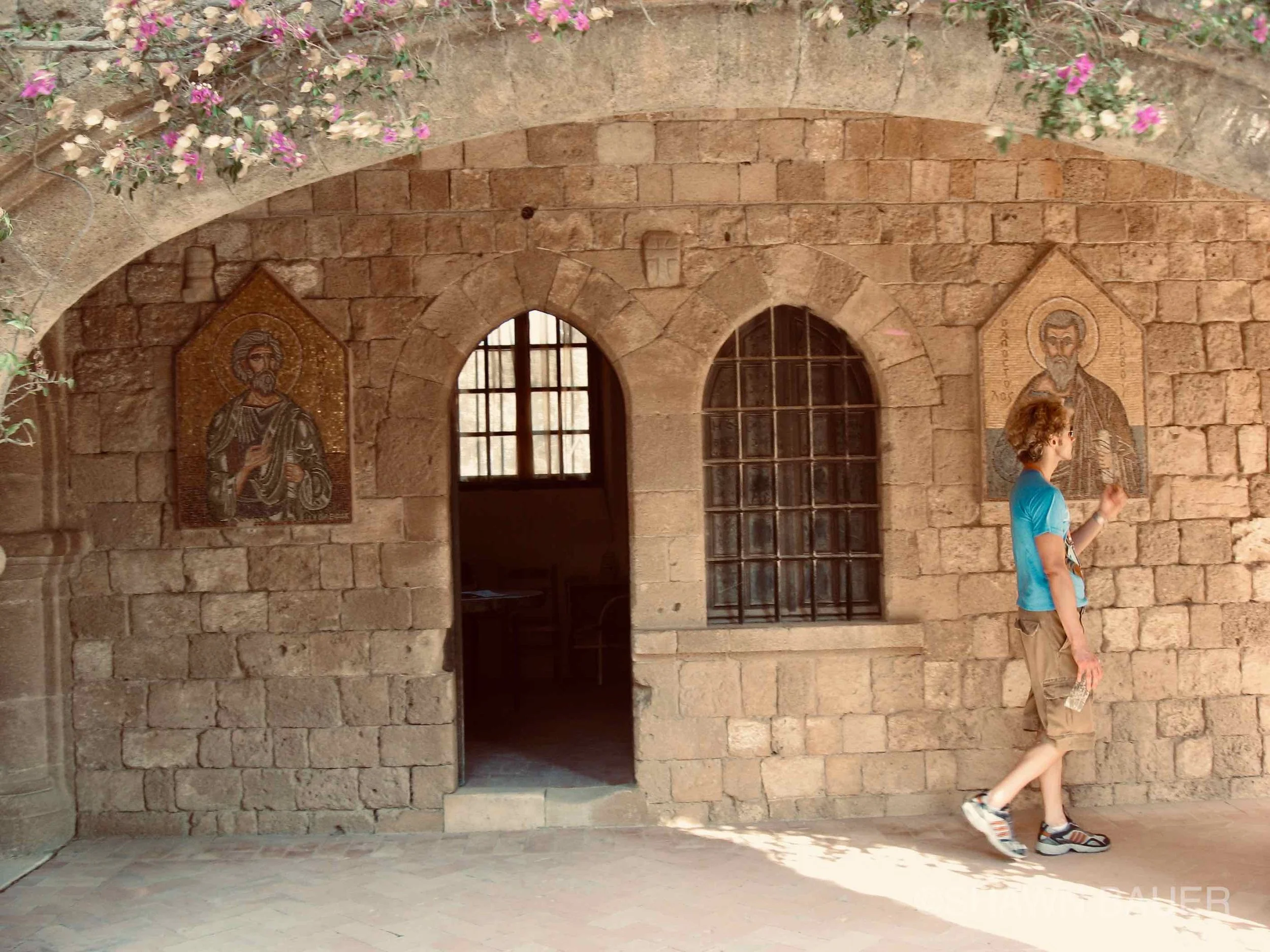 A person walking past a stone building with two stained glass icons of religious figures on its wall, arched windows, and pink flowering plants on the upper part of the wall. The person is wearing a blue shirt, beige shorts, and sneakers.