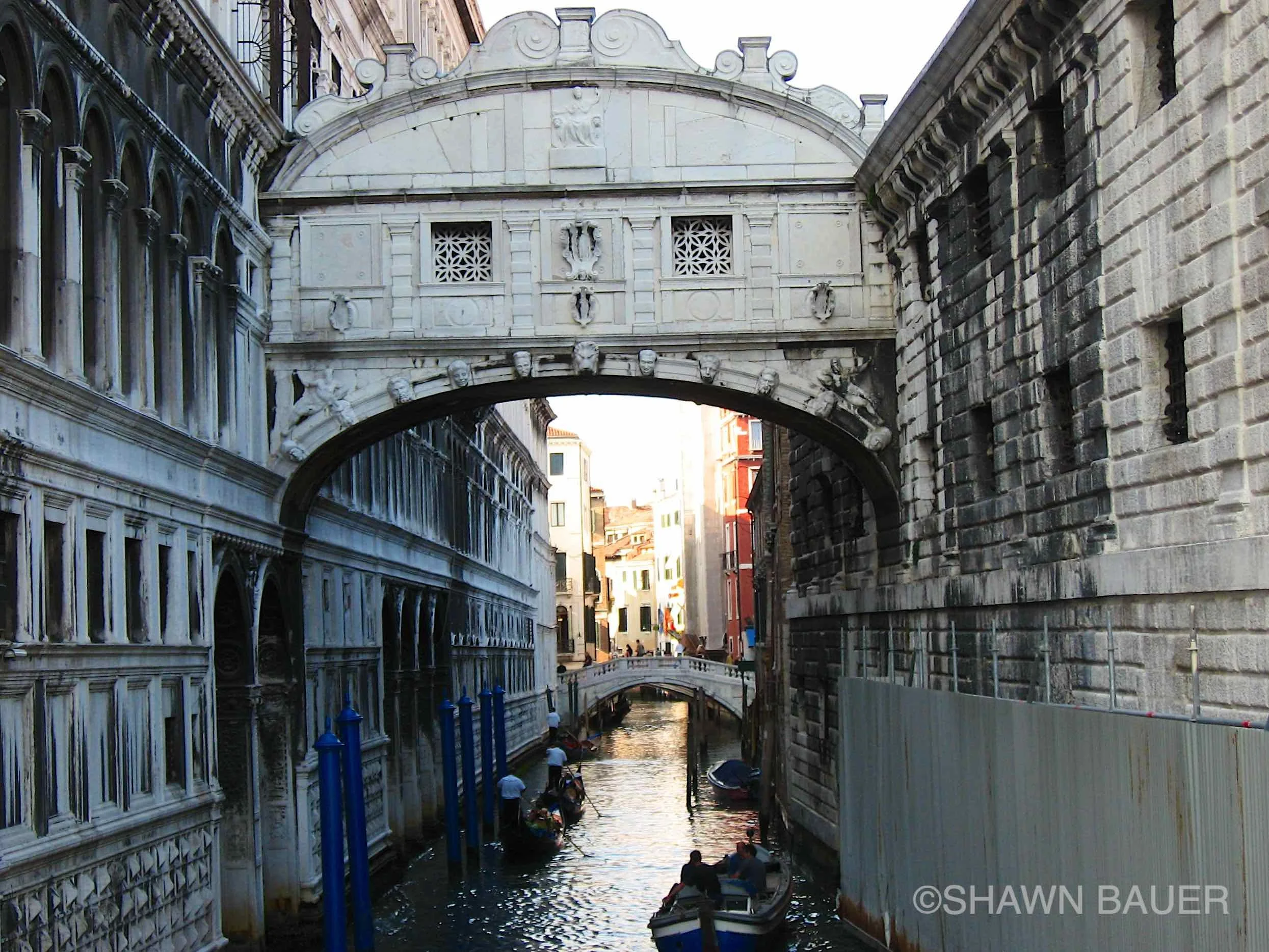 A scenic view of a narrow canal flanked by historic buildings on both sides, with children riding in boats and people walking along the waterway. An ornate white stone bridge with decorative details spans the canal.