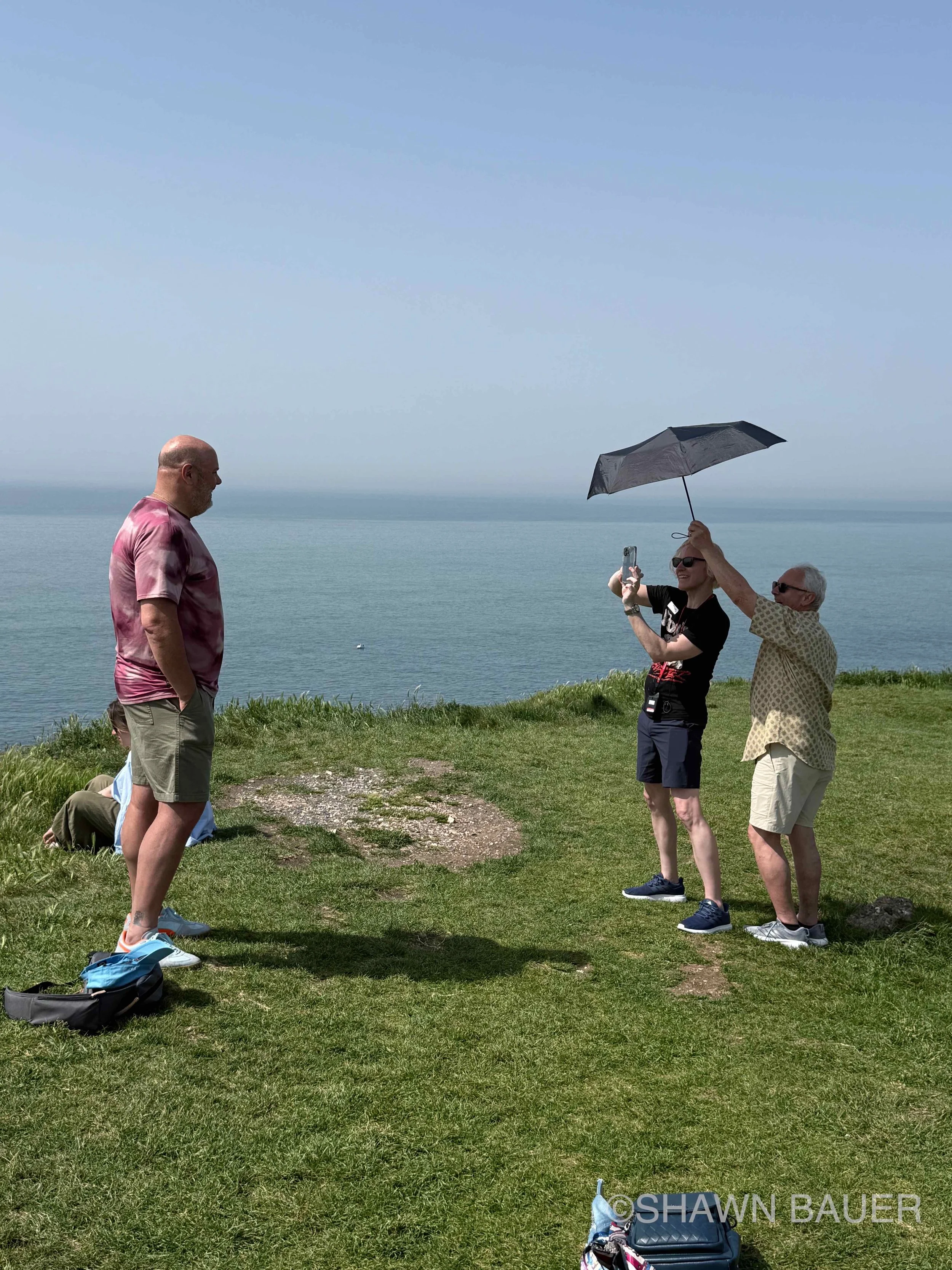Three people standing on a grassy area near the ocean with a cloudy sky, two of them are taking a photo of the third person.