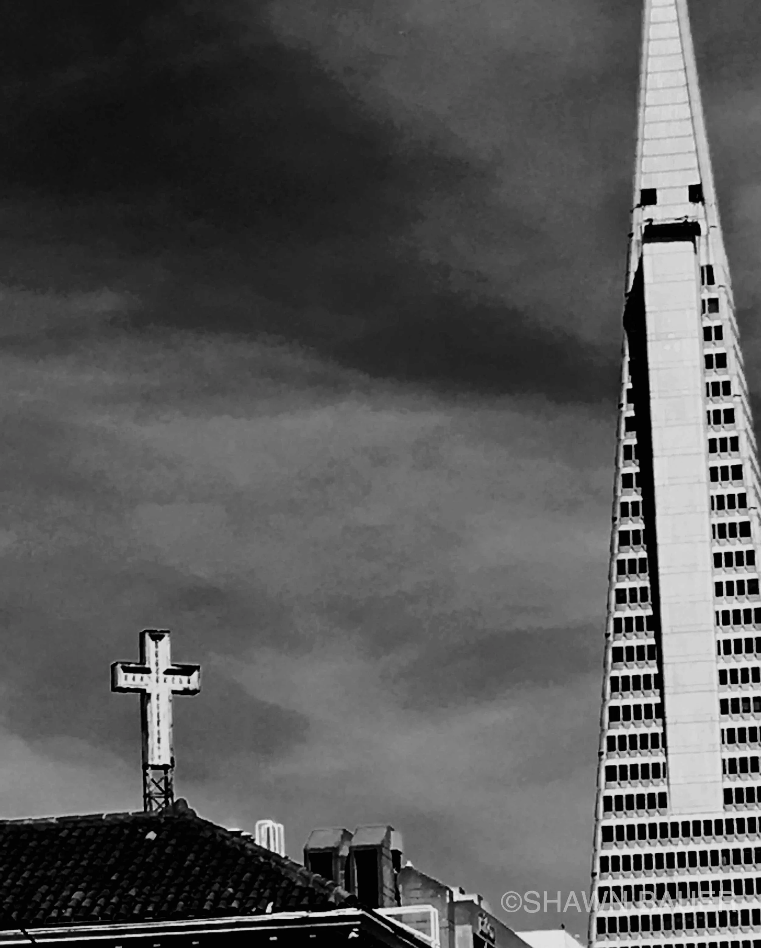 Black and white photo of city skyline including a tall skyscraper with a pointed top, a cross-shaped structure, and various rooftops against a cloudy sky.