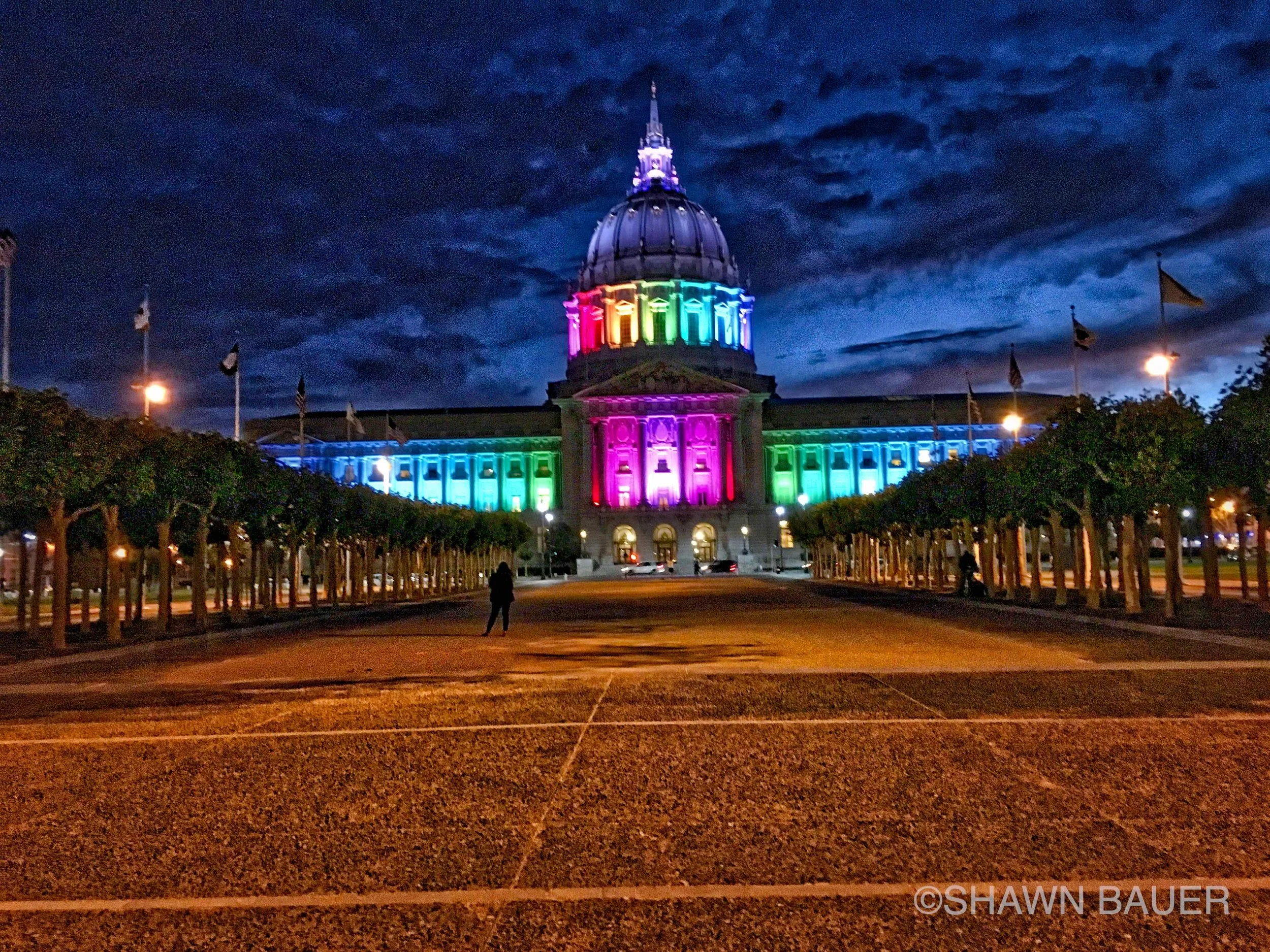 The Texas State Capitol building illuminated with rainbow colors at night, with a cloudy sky overhead, trees lining the area, and a few people walking nearby.