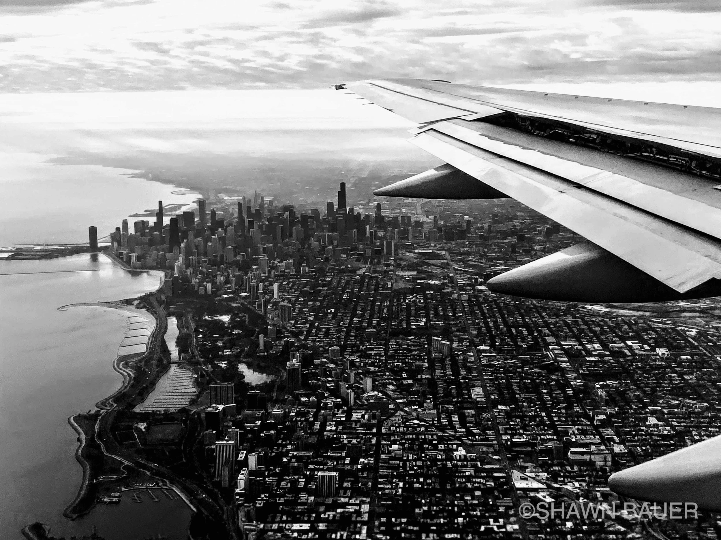 Black and white aerial photograph of a cityscape with numerous high-rise buildings, a body of water along the city, and airplane wing visible in the upper right corner.