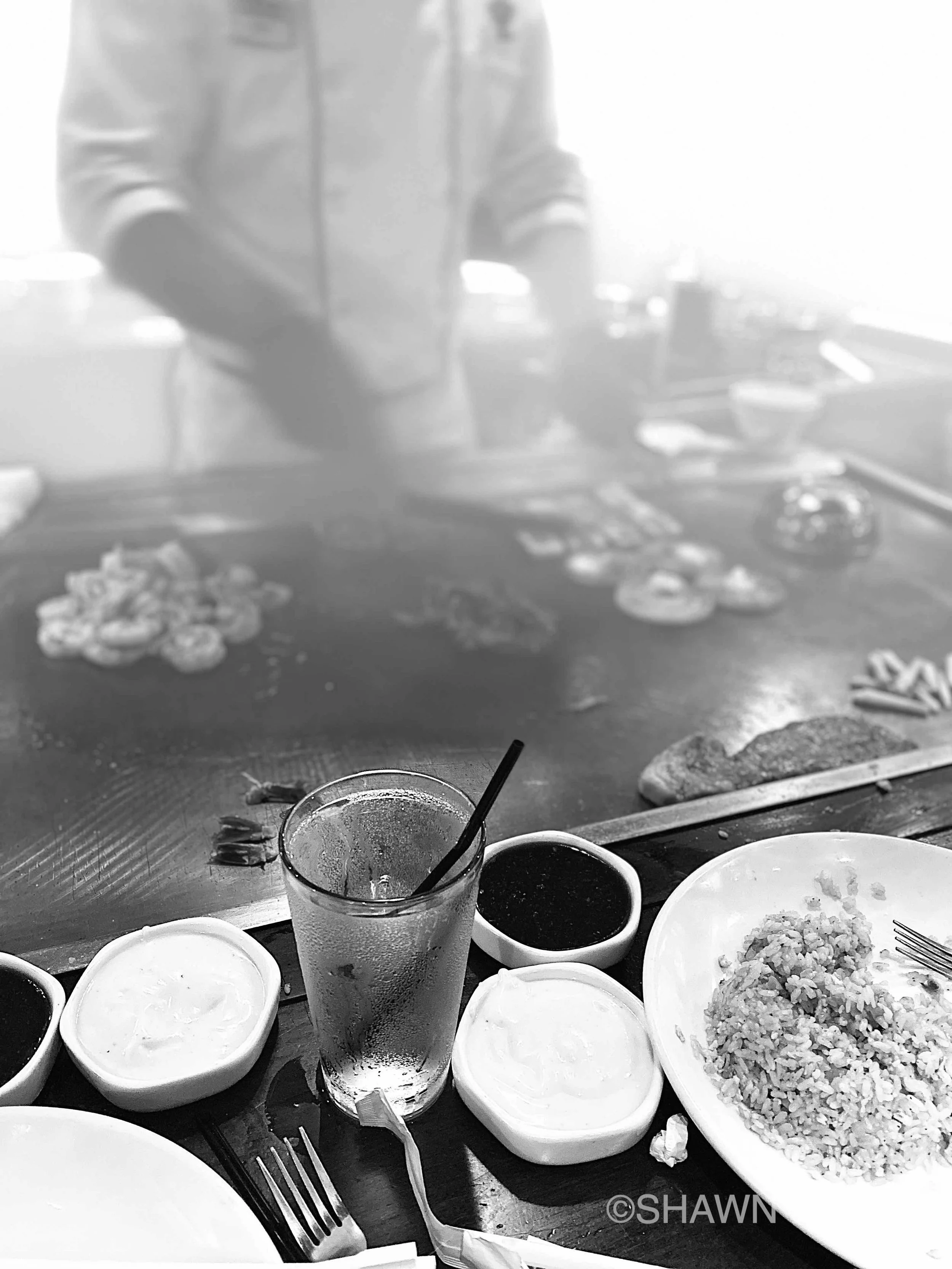 A table filled with various bowls of food, cups with beverages, utensils, and a plate of rice, indicating a meal setting at a restaurant or home. The background shows a person's torso and hands, suggesting they are preparing or serving food.