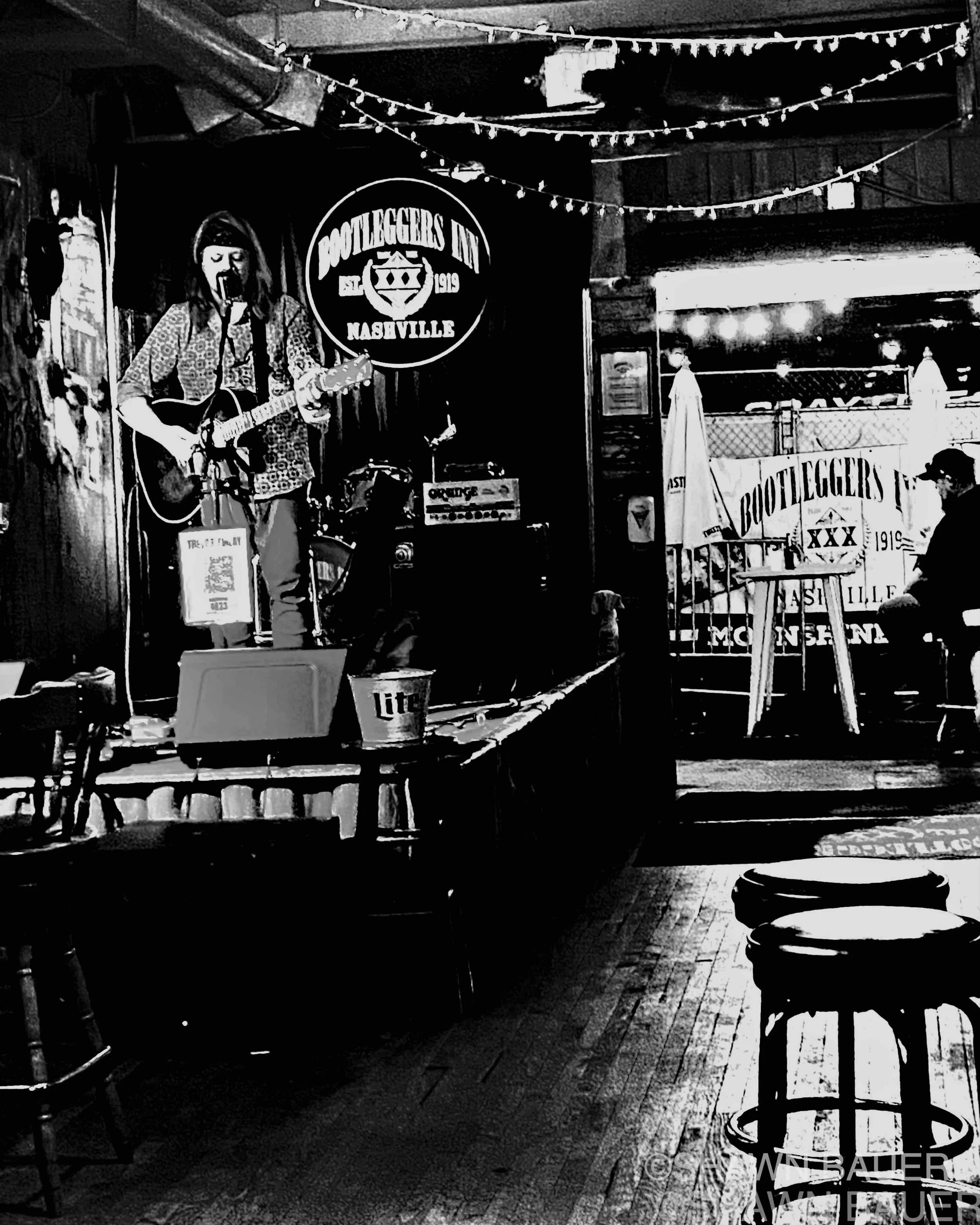 A woman performing on a stage at a Nashville venue called Bootleggers Inn. She is playing a guitar and singing into a microphone, with a performer sign and string lights above her. Audience seating and a table with a Miller beer bucket are visible in the foreground.