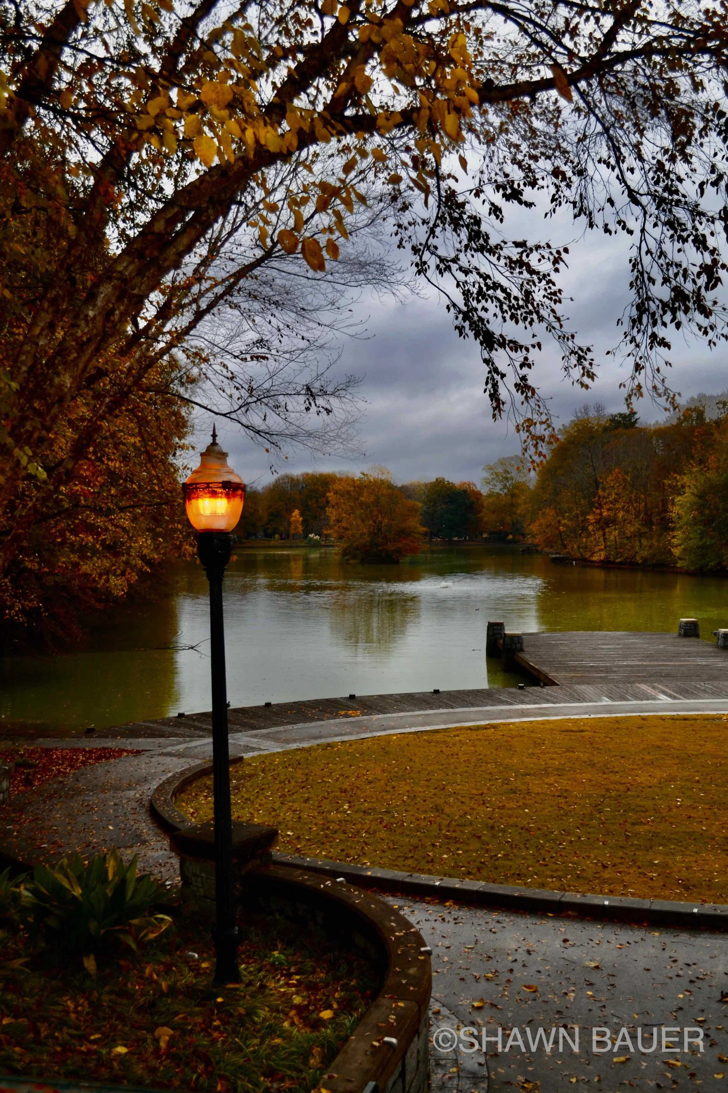 A lakeside park scene at dusk with a lit street lamp, autumn trees with yellow and orange leaves, a wooden dock extending into a calm lake, and cloudy sky overhead.