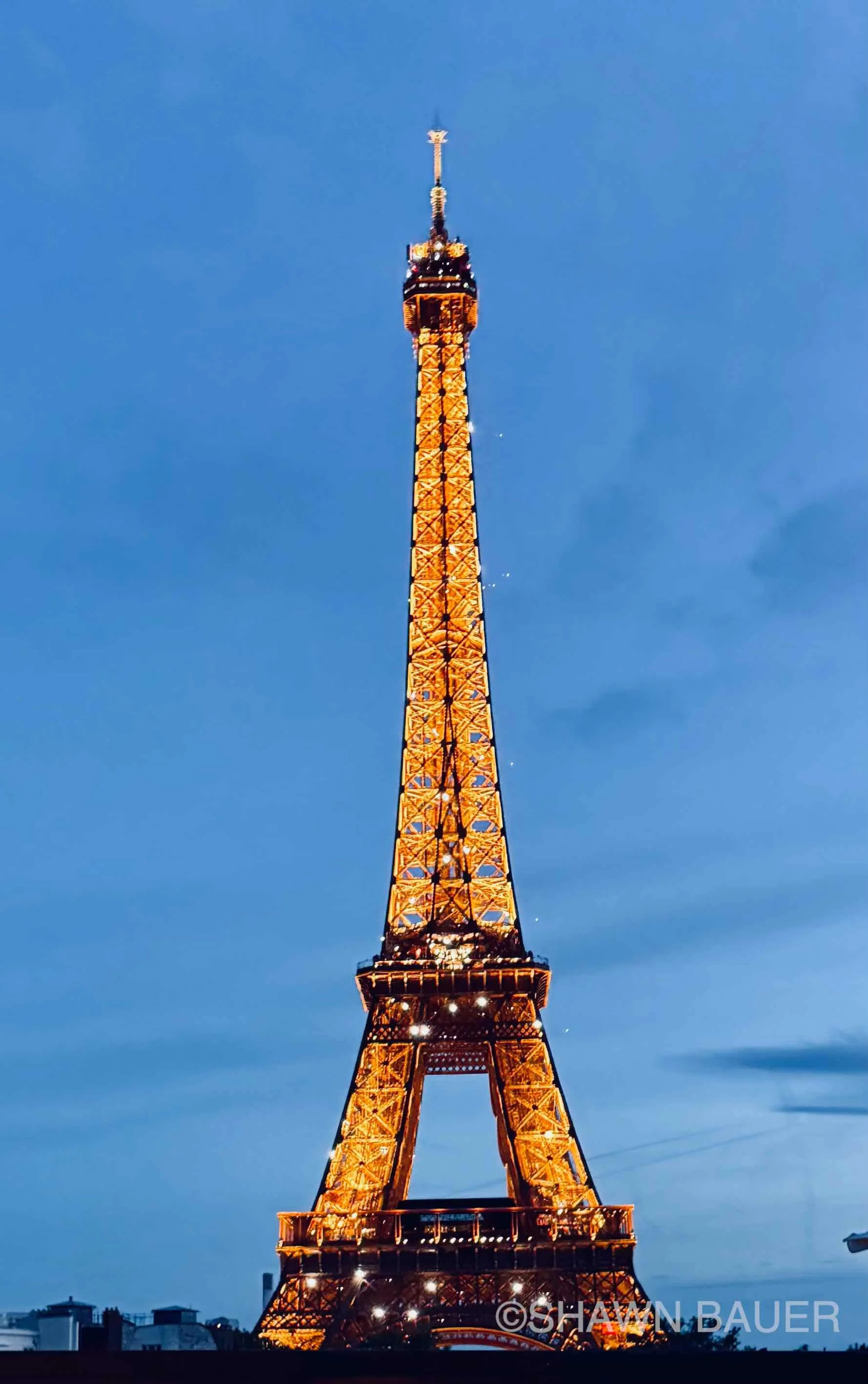 The Eiffel Tower lit up at night against a blue sky, with city buildings at the bottom.