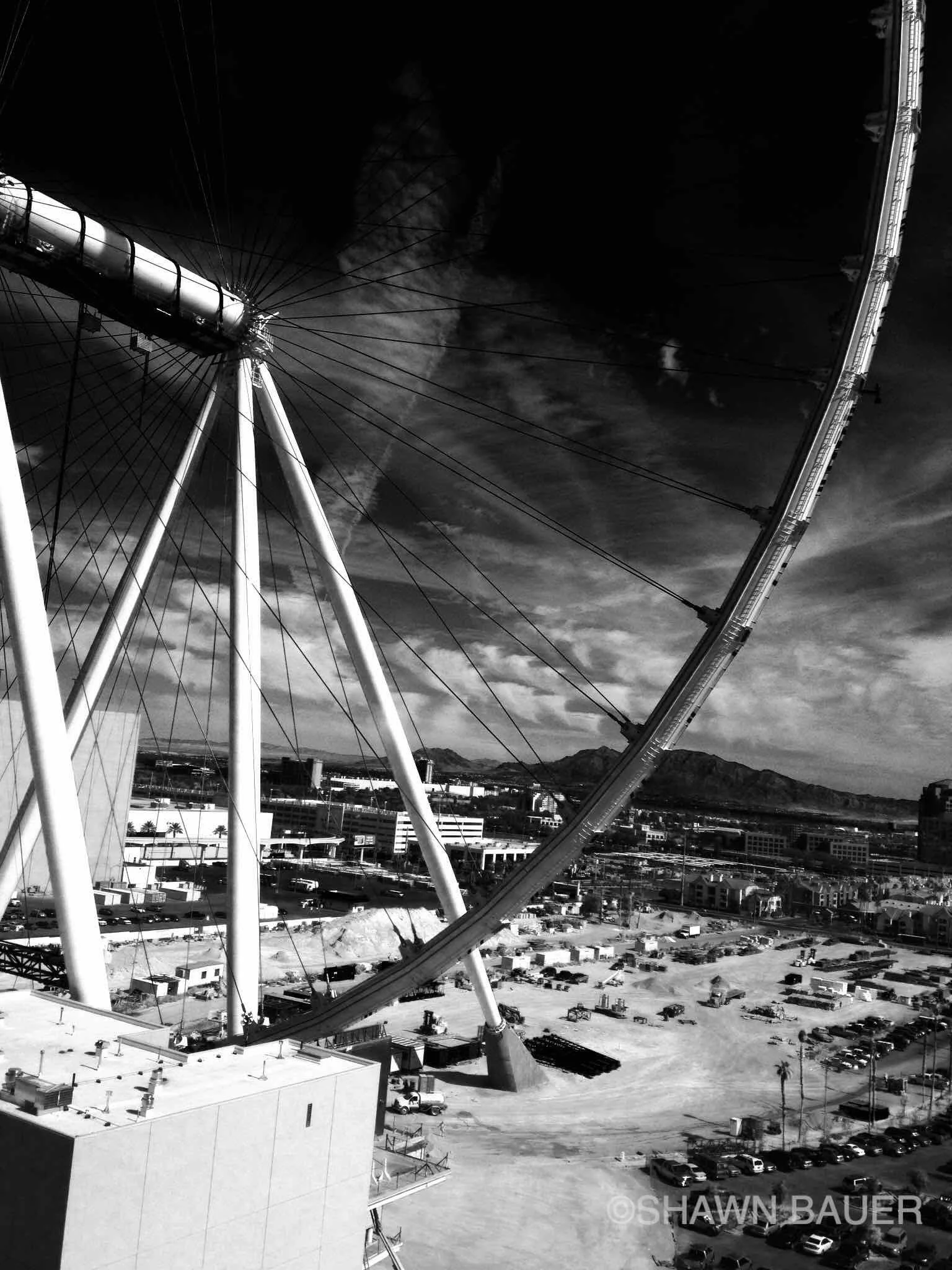 Black and white photo of a large Ferris wheel in an urban area with a cityscape and mountains in the background.