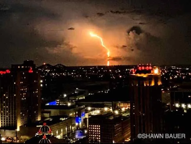 Lightning strikes over a city skyline at night with cloudy sky and illuminated buildings.