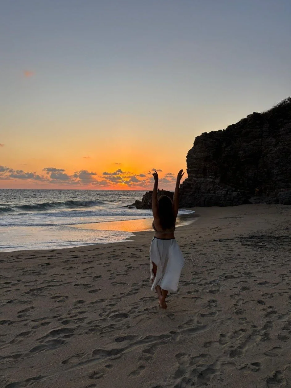 Frau, die am Strand bei Sonnenuntergang tanzt, mit erhobenen Armen, Sand und Felsen im Hintergrund. Sie geht ihren Lebensweg.