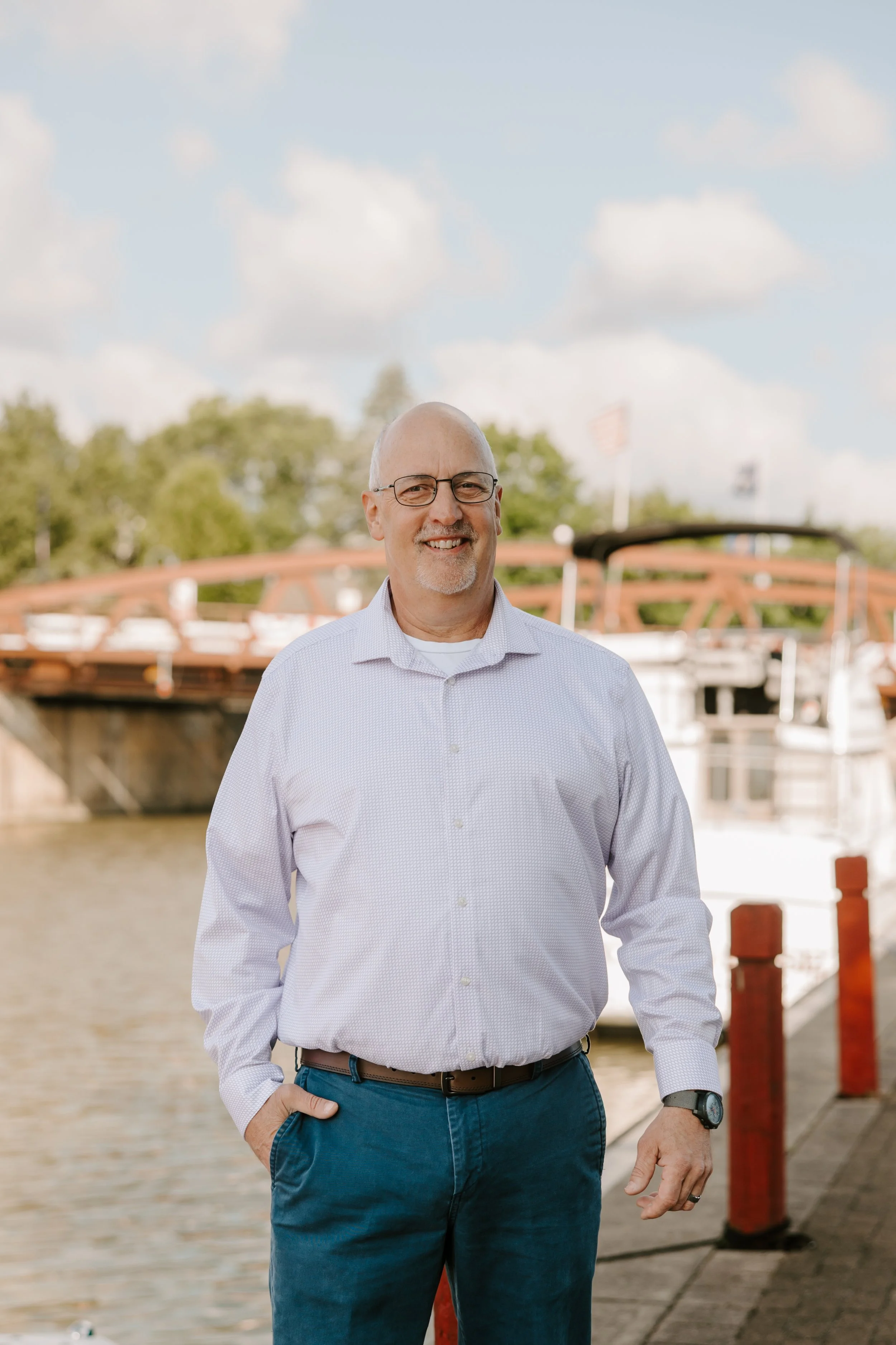 A middle-aged man with glasses, a white shirt, and blue pants standing on a dock by a body of water, with boats and trees in the background.
